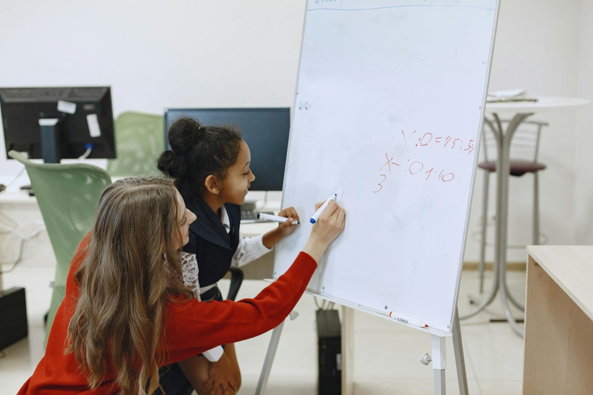 A teacher helps a young student solve maths equations on a whiteboard