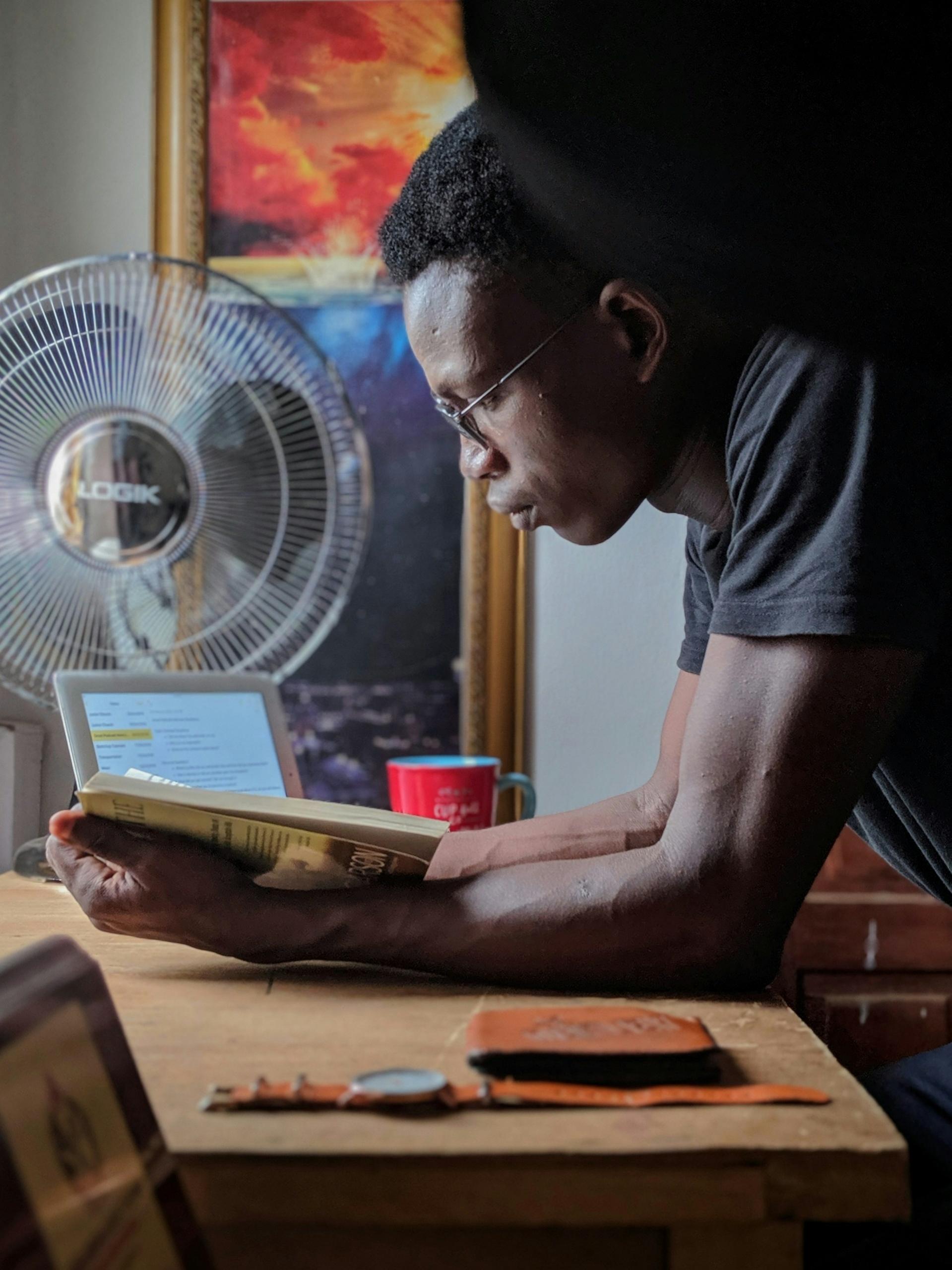 A man wearing glasses and a blue shirt reads a book while leaning on a table. 