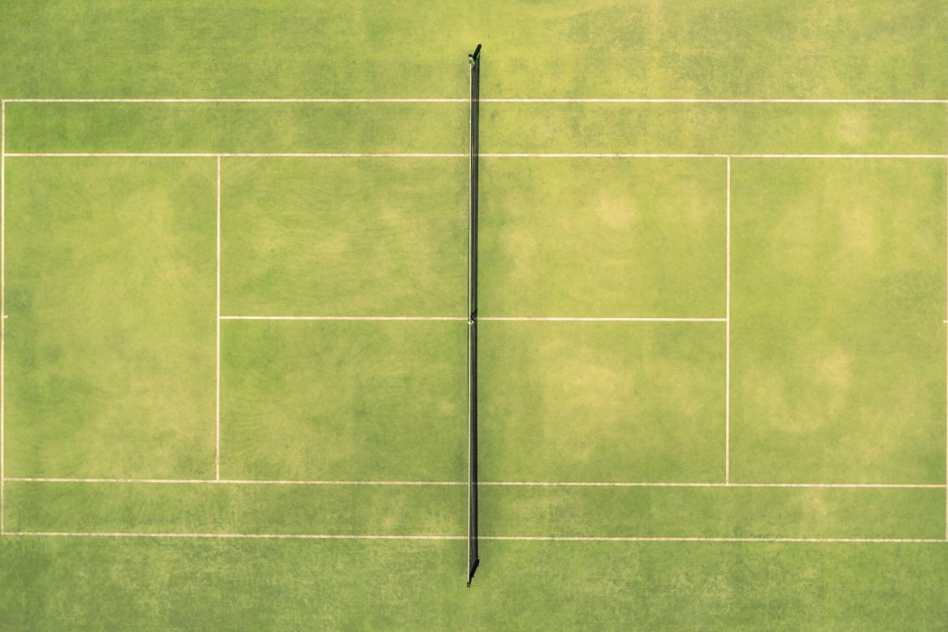 A tennis court viewed from above with the net running down the centre and white court lines visible on the grass.