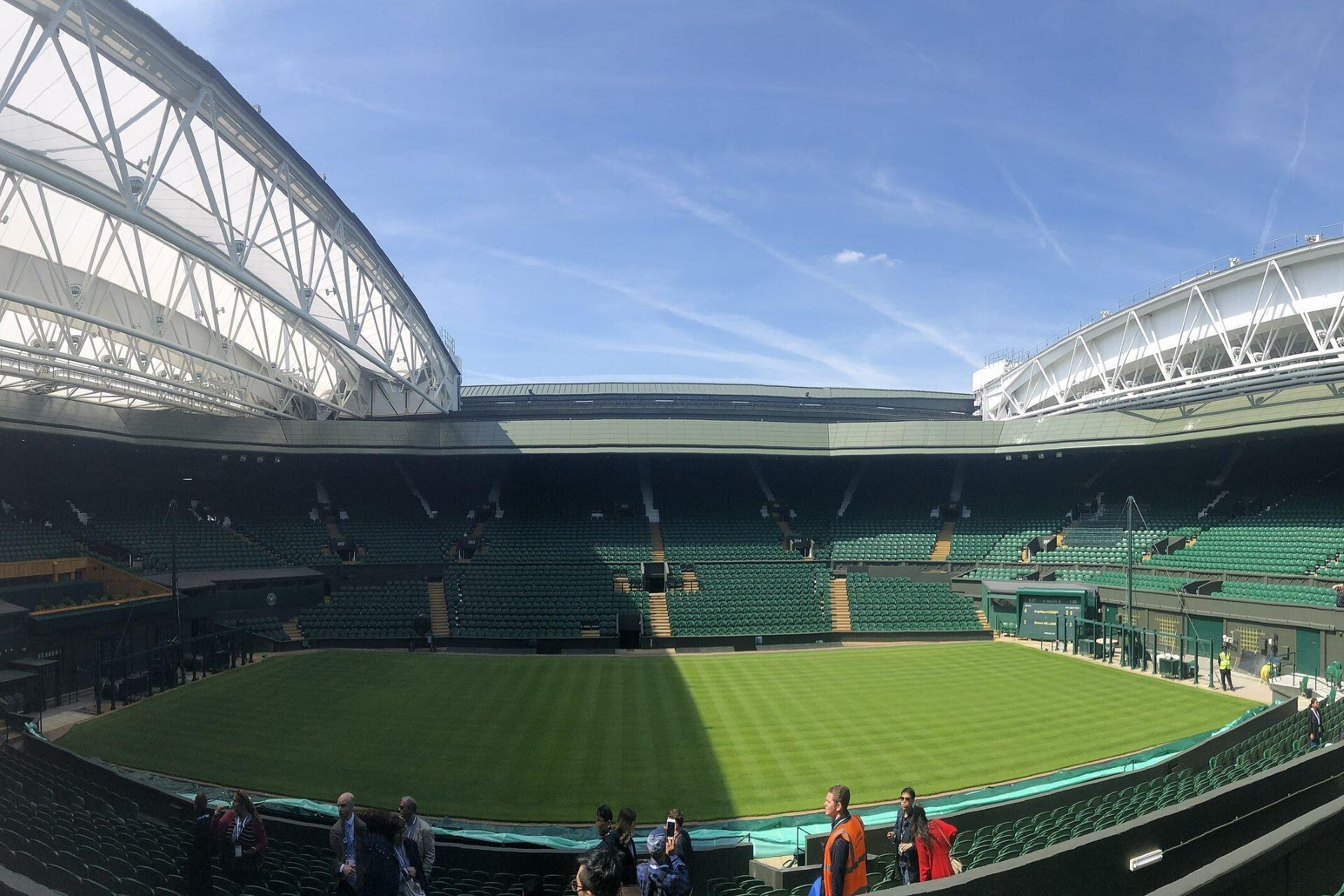 Wimbledon Centre Court with green grass, rows of seats, and the open roof structure under a blue sky.
