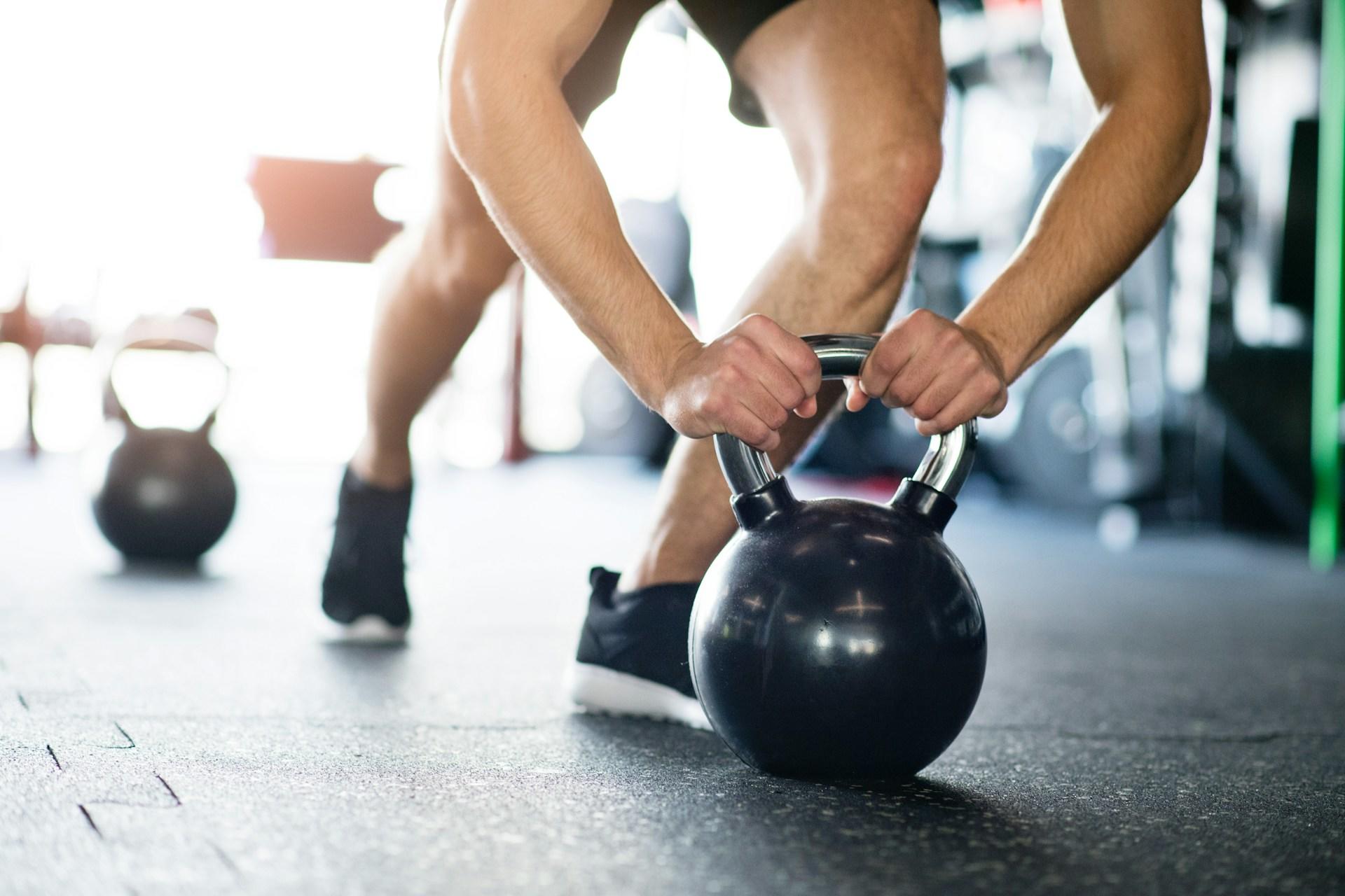A person lunges forward to grasp a kettlebell with two hands in a gym.