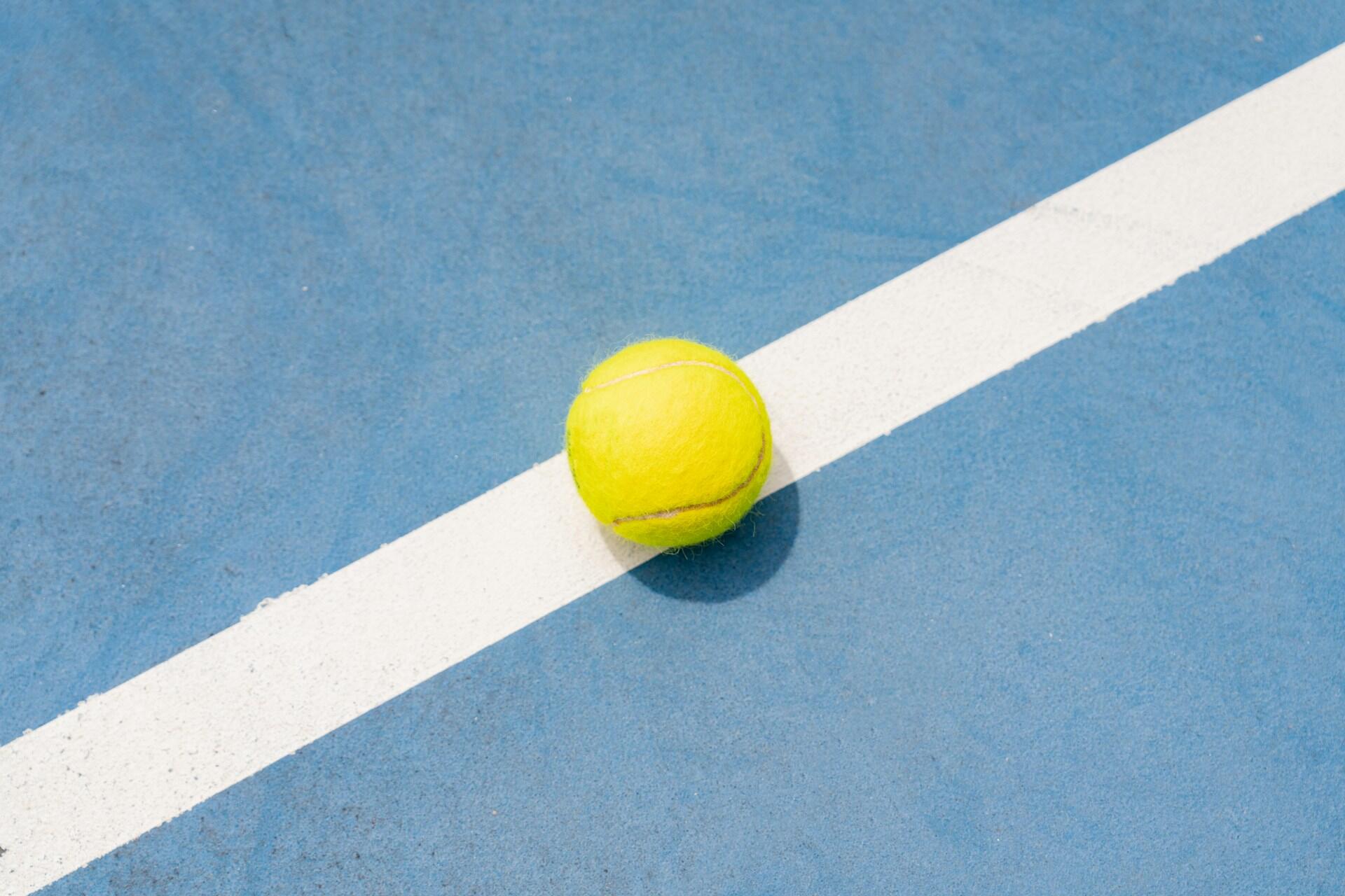 A yellow tennis ball resting on a white line on a blue tennis court surface.