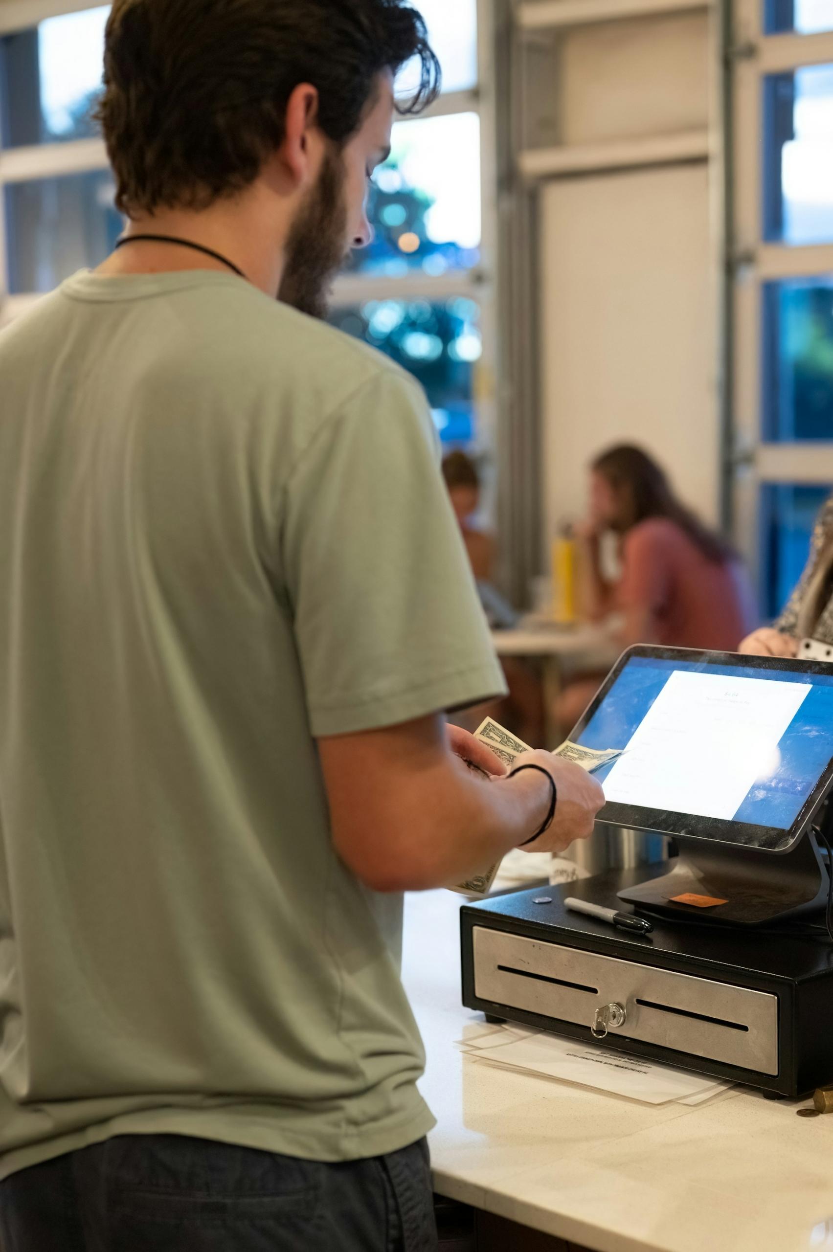 A man in a green shirt stands behind a cash register with money in his hands.
