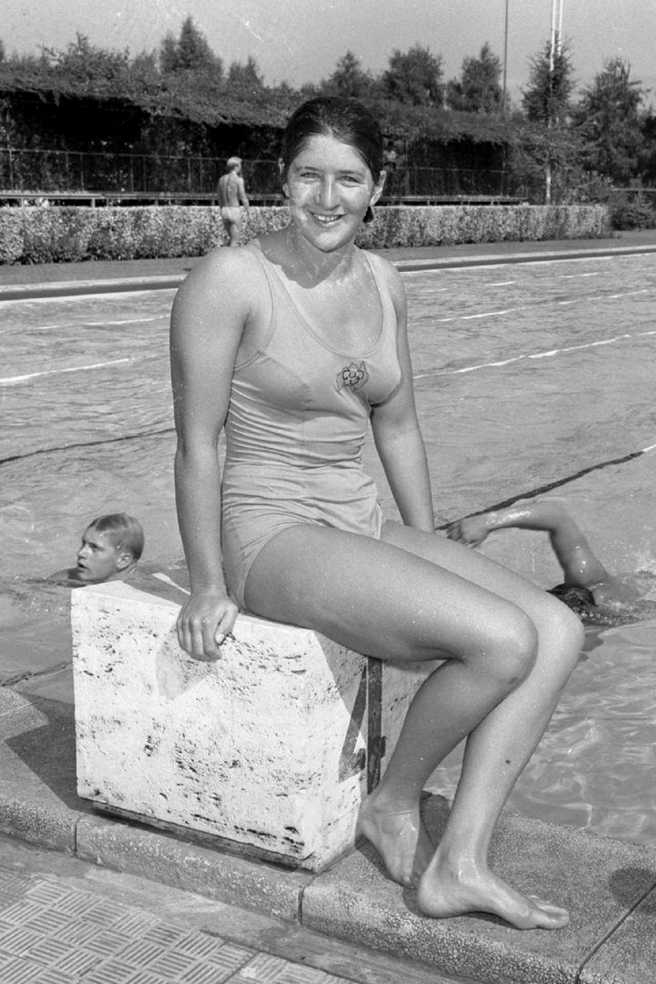 A woman sits on a dive block by a swimming pool.