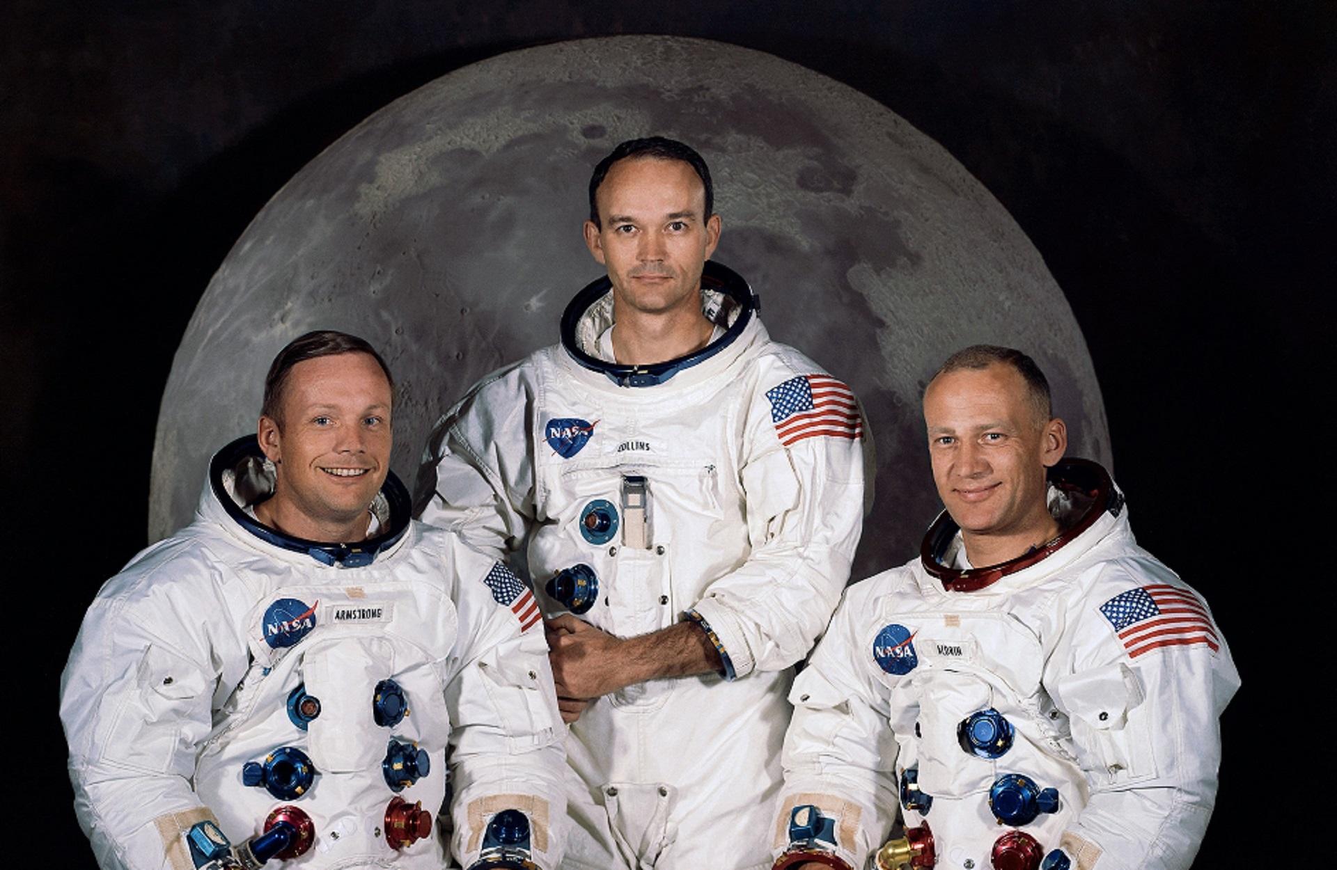 Three men in space suits, minus helmets, posed against a background of the full moon. 