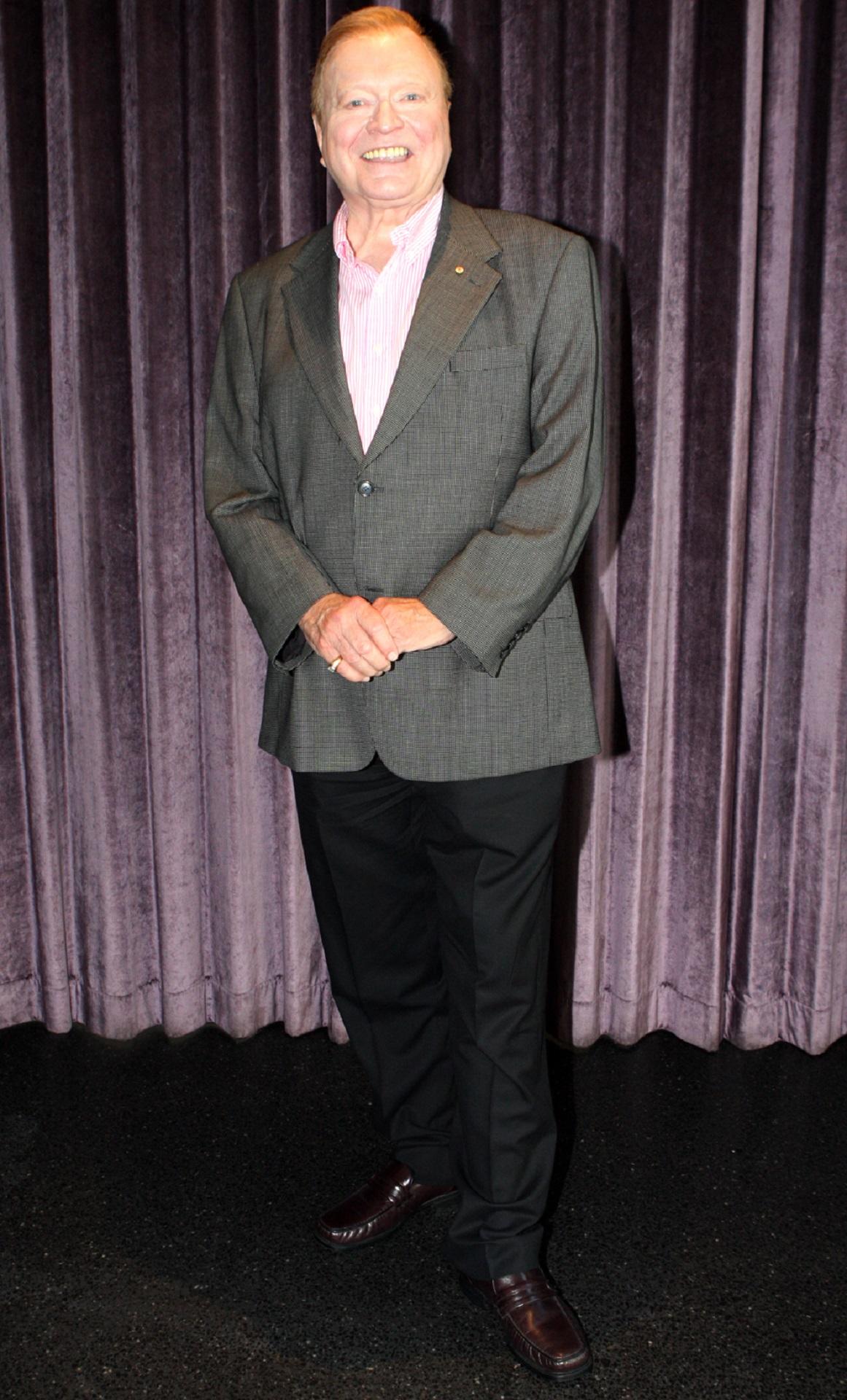 A man in a grey blazer stands in front of a purple curtain. 