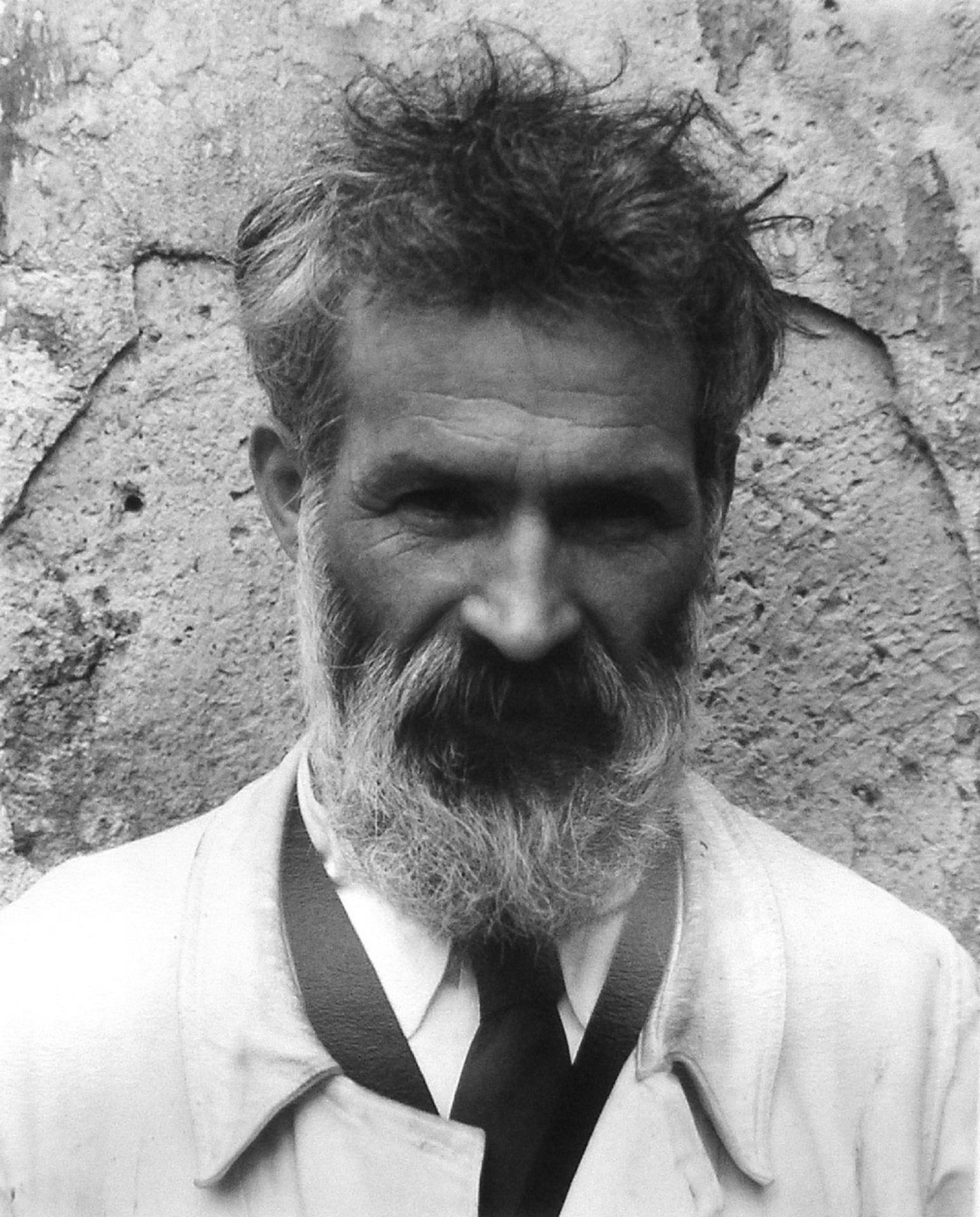 A greyscale of a bearded man wearing a suit and tie under a light overcoat.