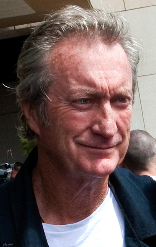 A grey-haired man wearing a white tee shirt under a dark collared shirt.
