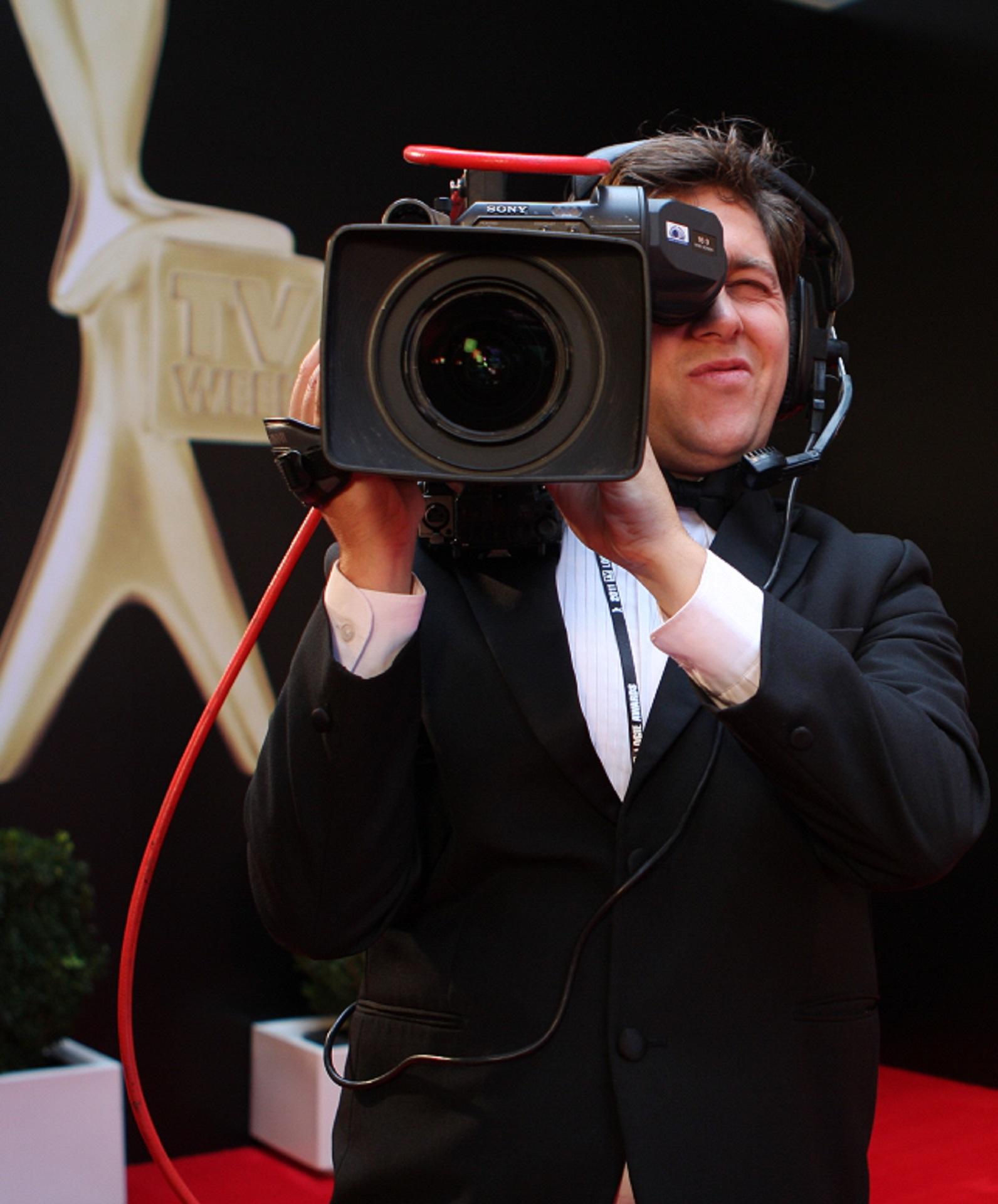 A man in a suit holds a TV camera with a Logie statue in the background.