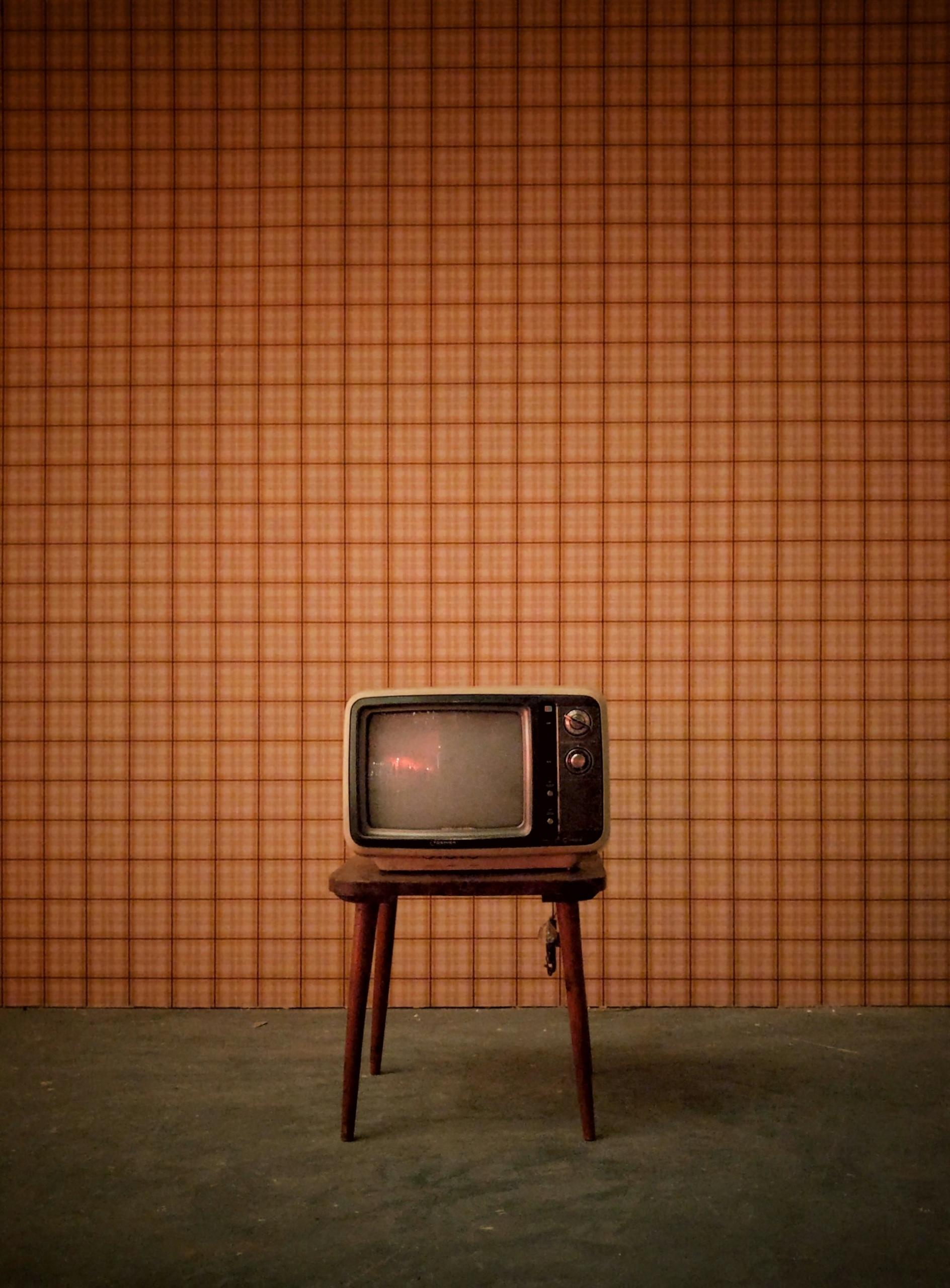 An old television on a spindly legged table in front of a brown grid wall. 