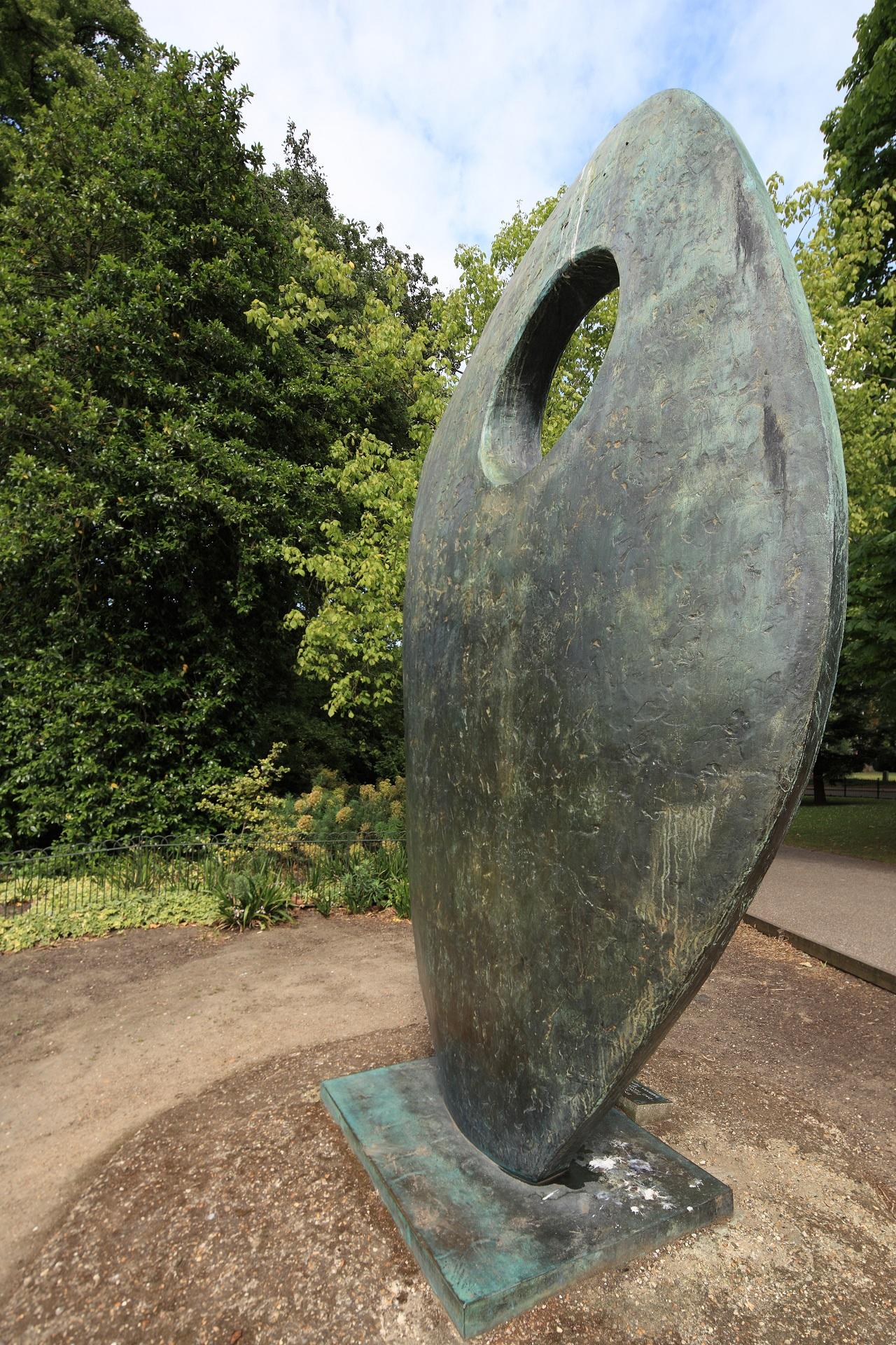 A grey sculpture stands in a clearing with greenery around it.