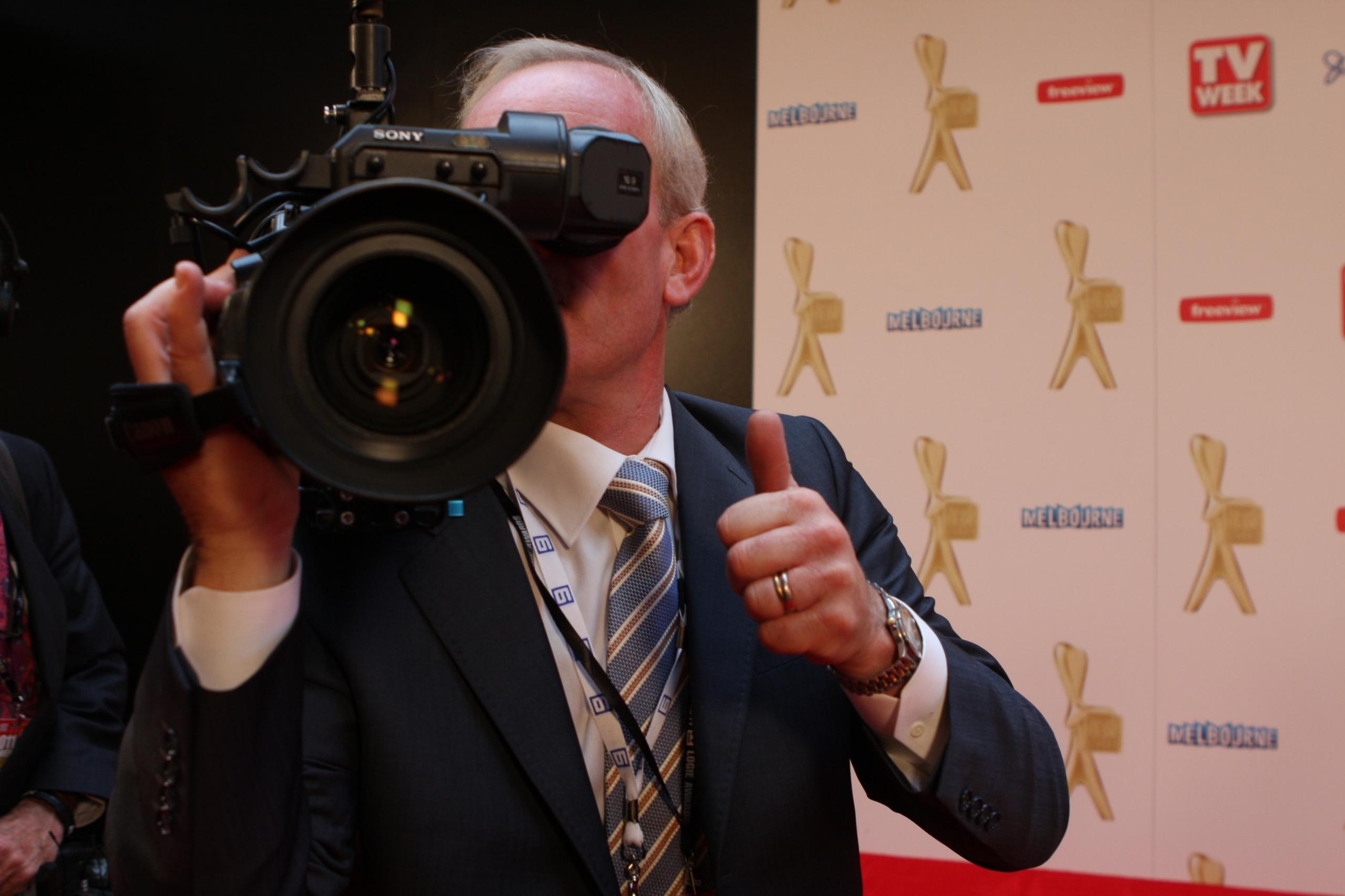 A man in a suit stands behind a camera in front of a promo wall.