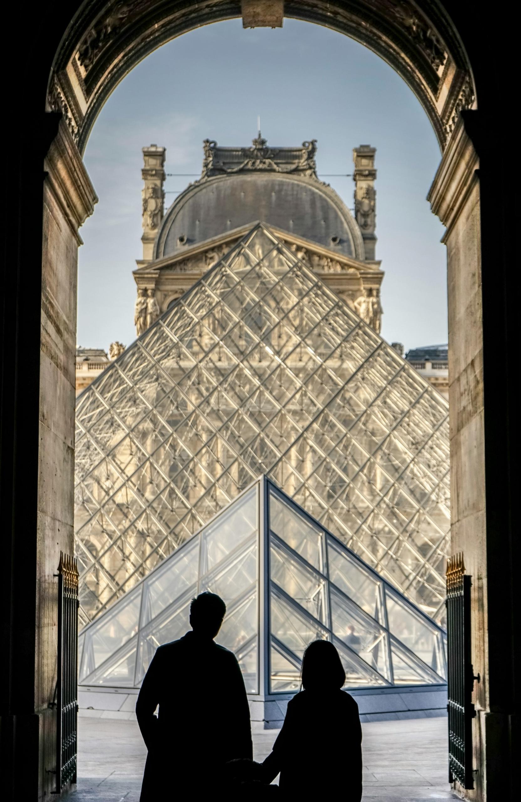 Two people in silhouette, framed in an archway, in front of a glass pyramid.