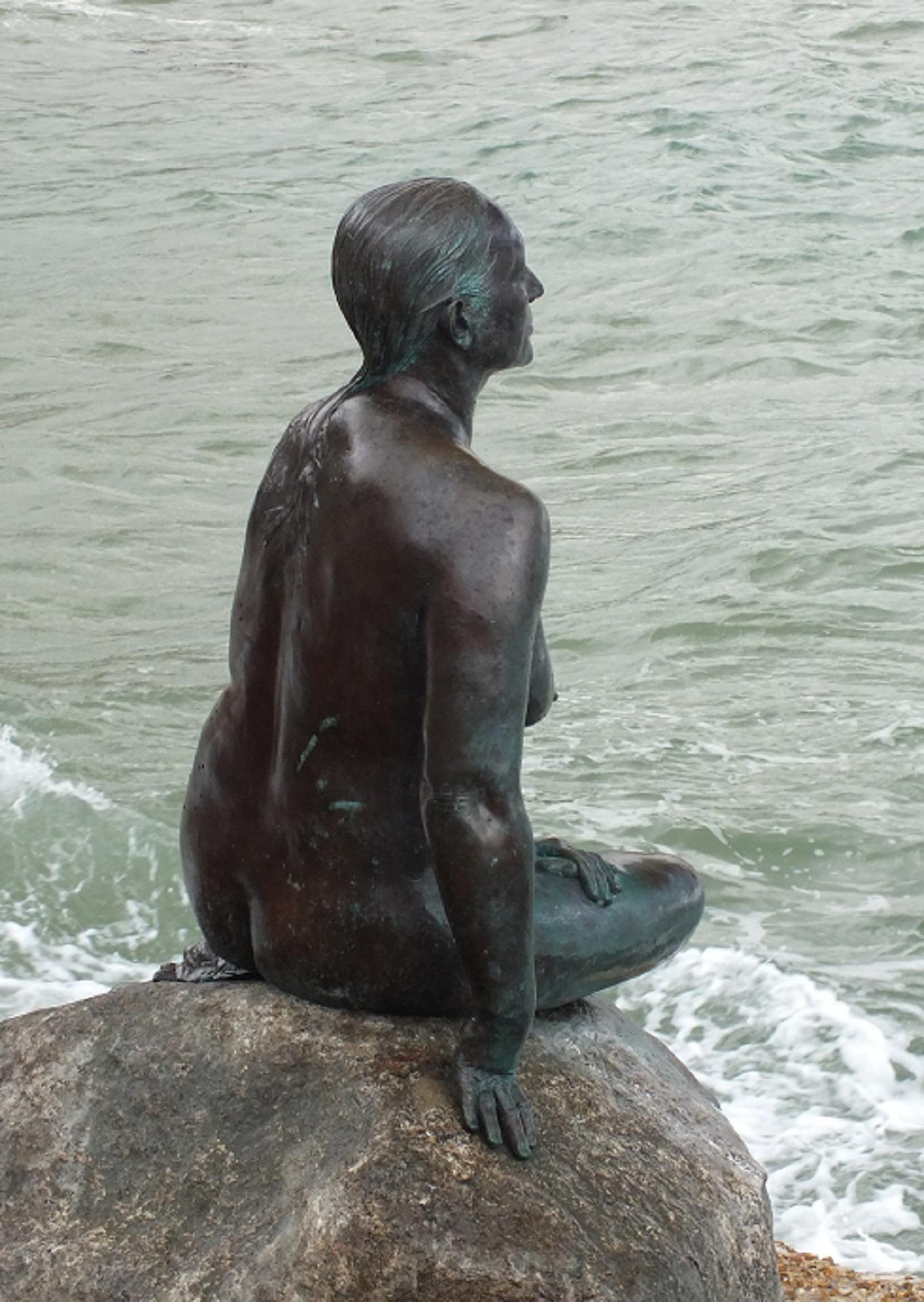 A bronze statue perched on a stone near the water. 