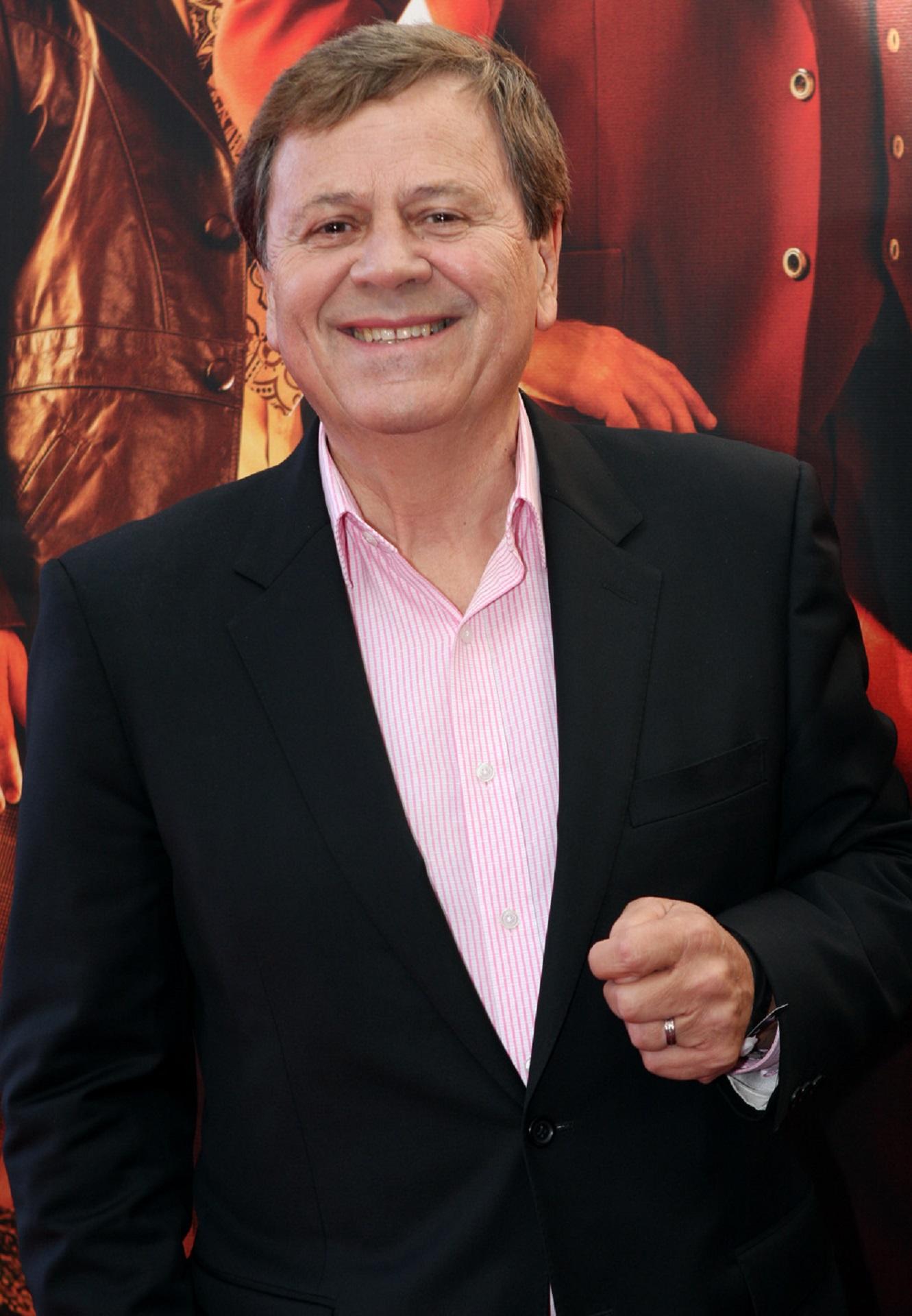 A smiling man wears a pink shirt and a black blazer. 