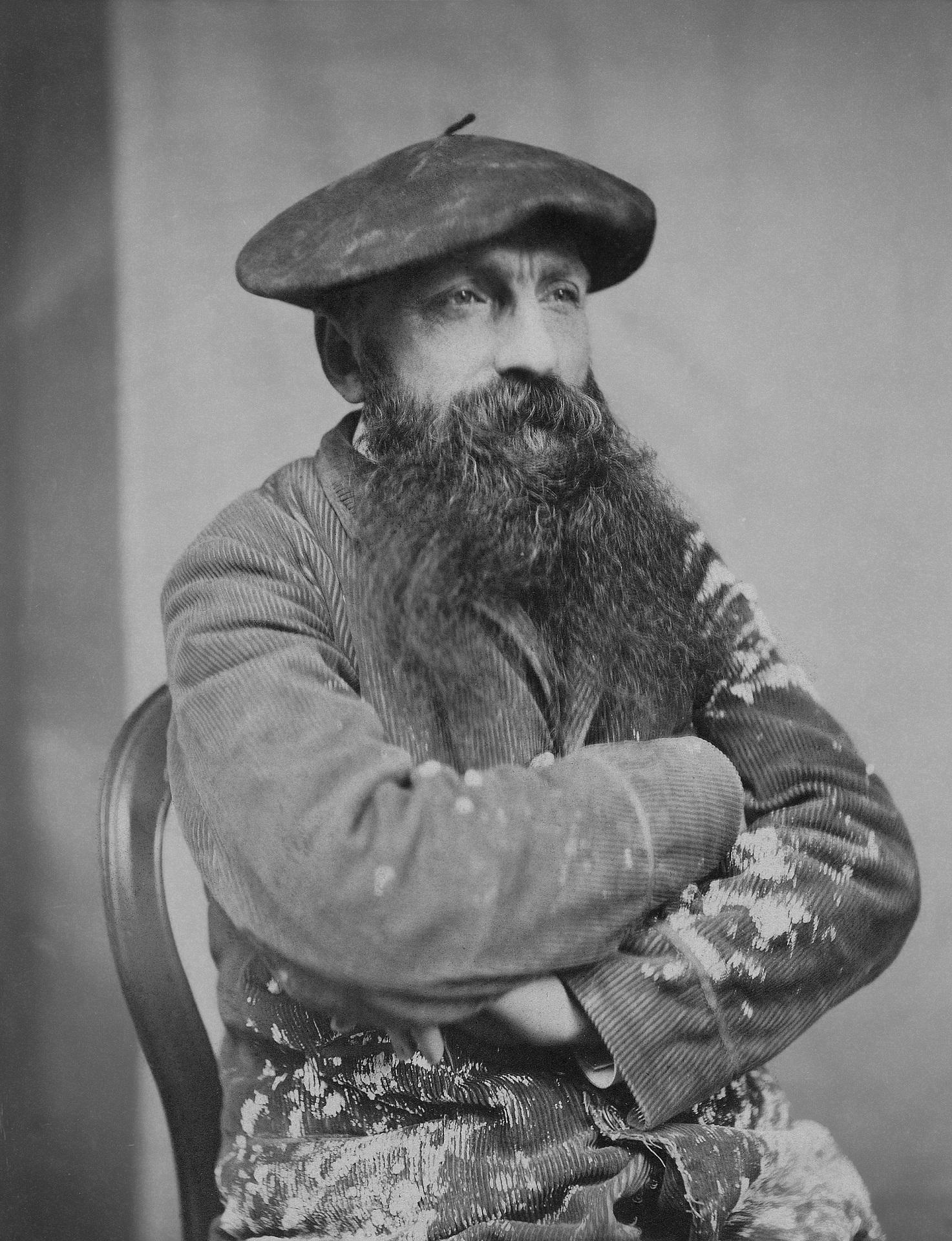 A greyscale image of a bearded man wearing a beret and sitting his his arms crossed.