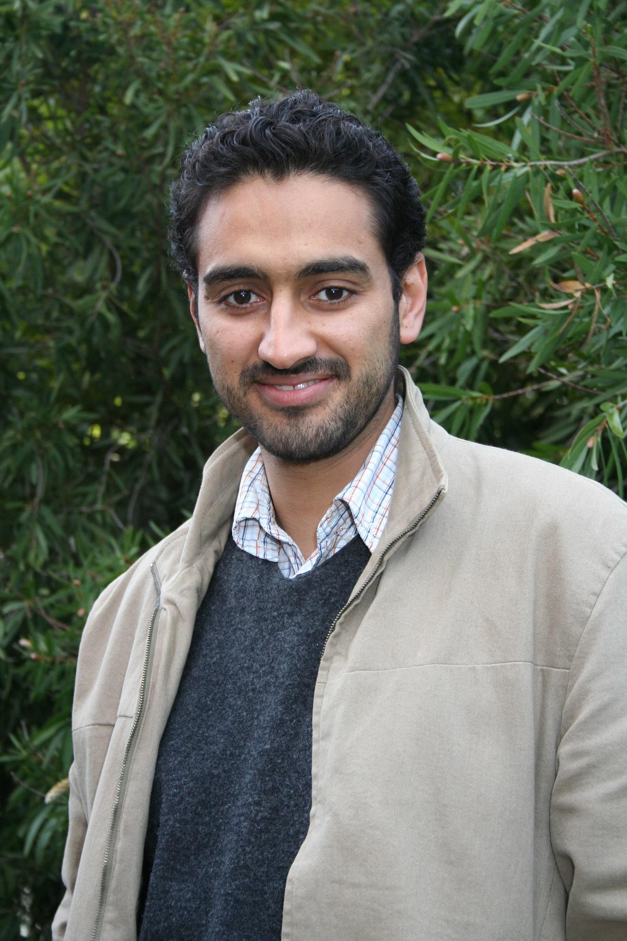 A smiling man wearing a tan jacket over a grey jumper and white shirt.