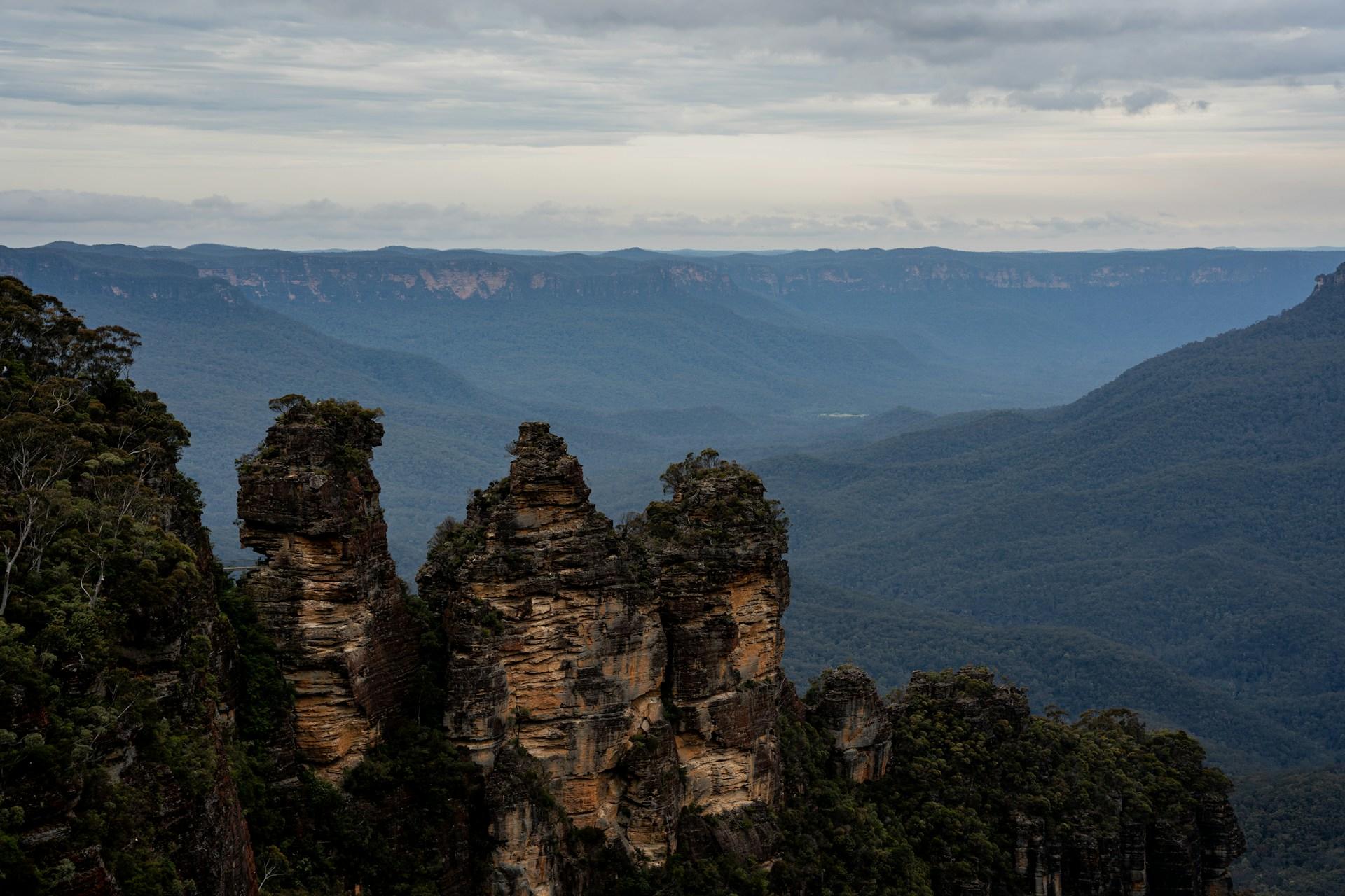 Three large rock formations in the foreground with a vast forested valley and mountain ranges in the background.