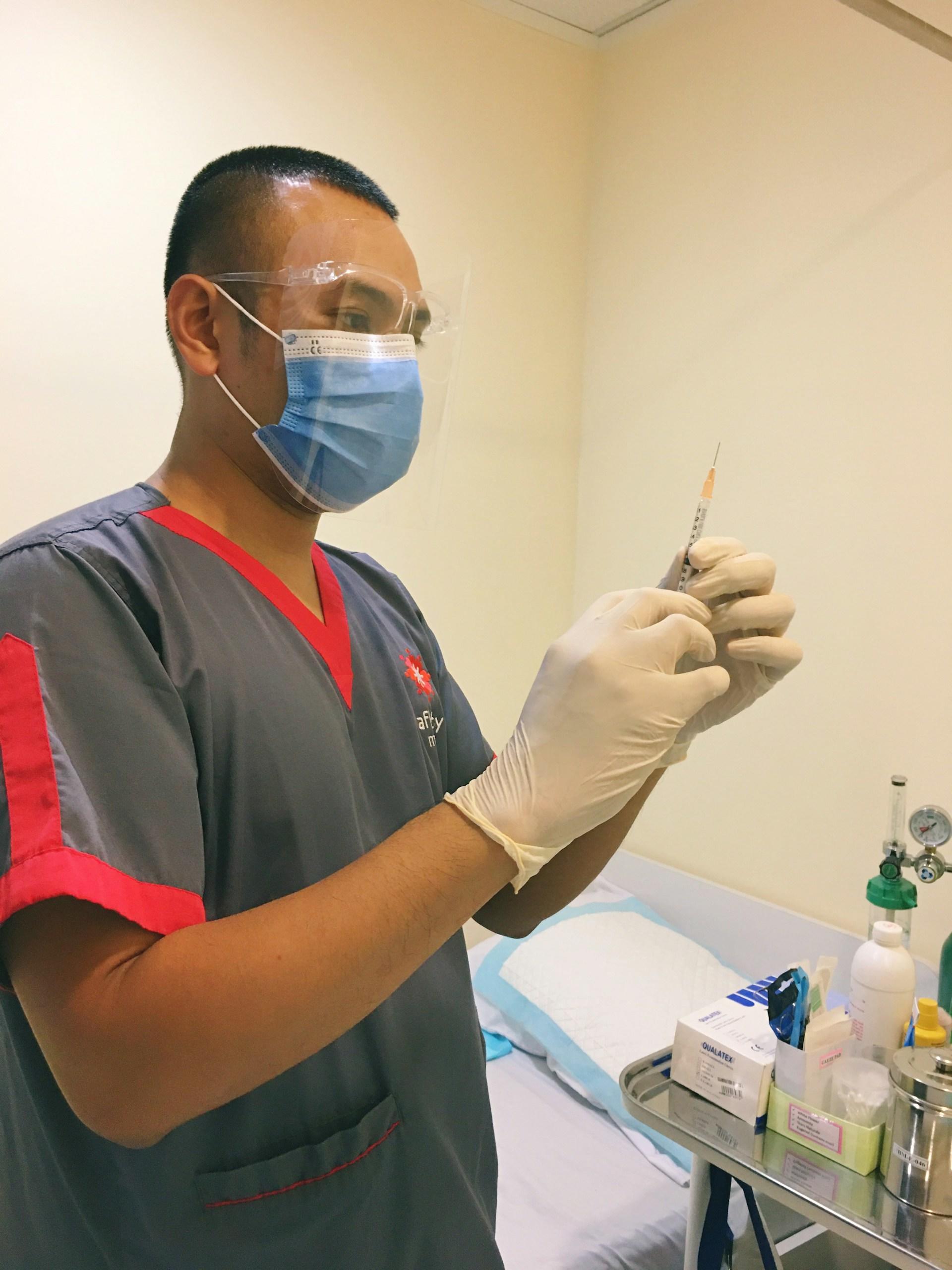 A man in a grey and red top with a face mask preparing an injection. 