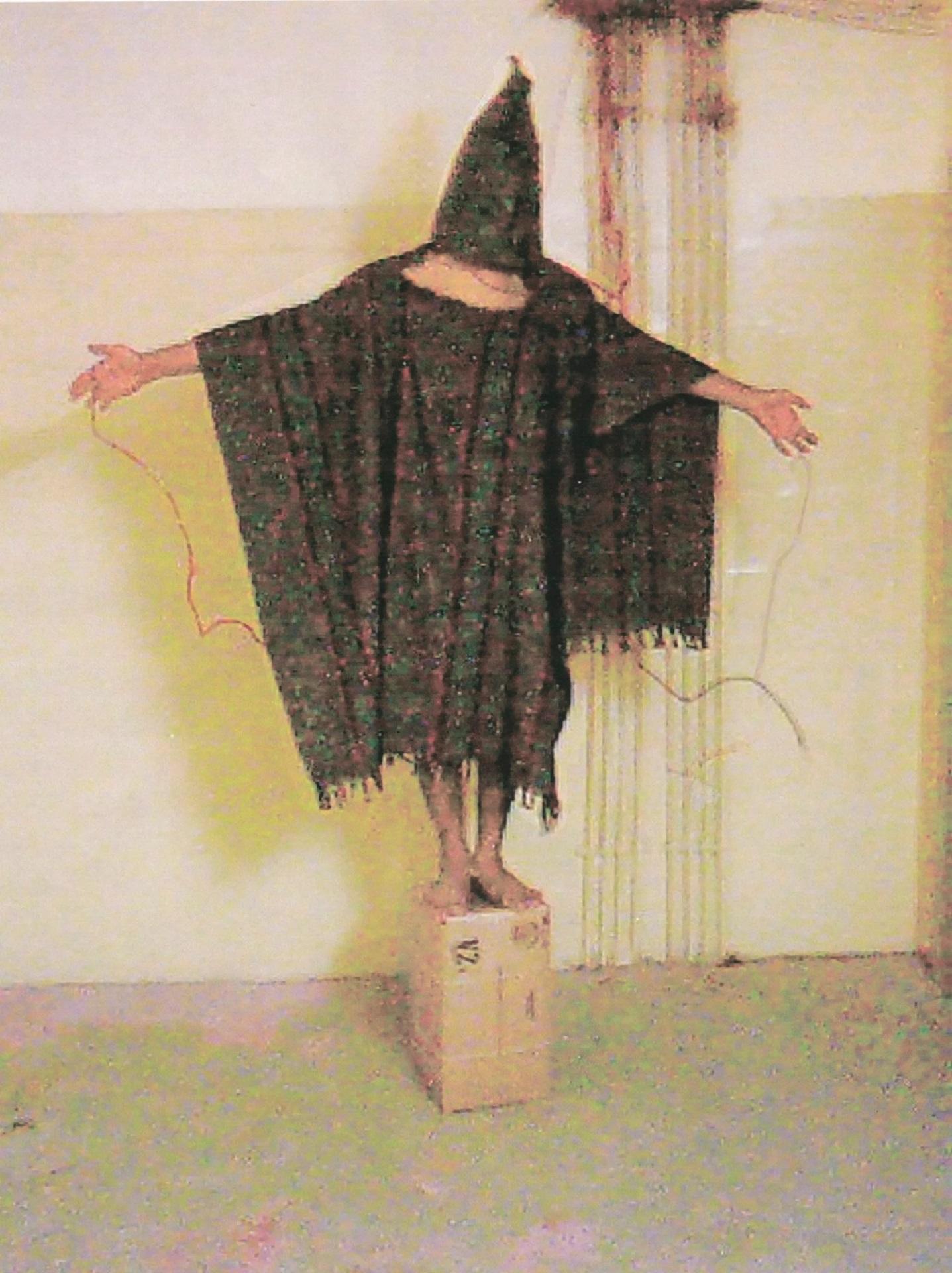 A body wearing a black poncho and hood stands on a box with arms outstretched.
