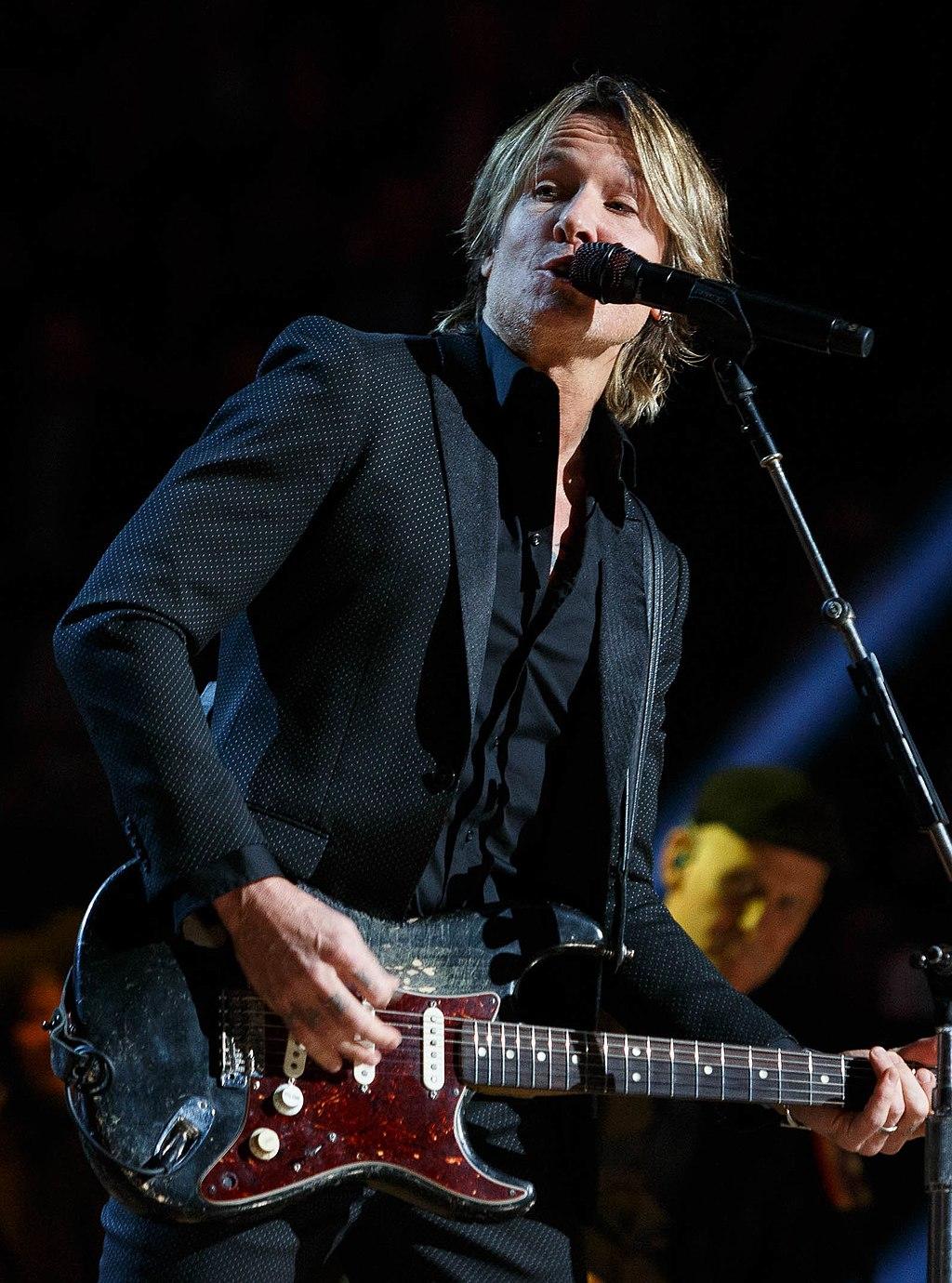 Keith Urban on stage playing guitar and singing, wearing a black dress shirt
