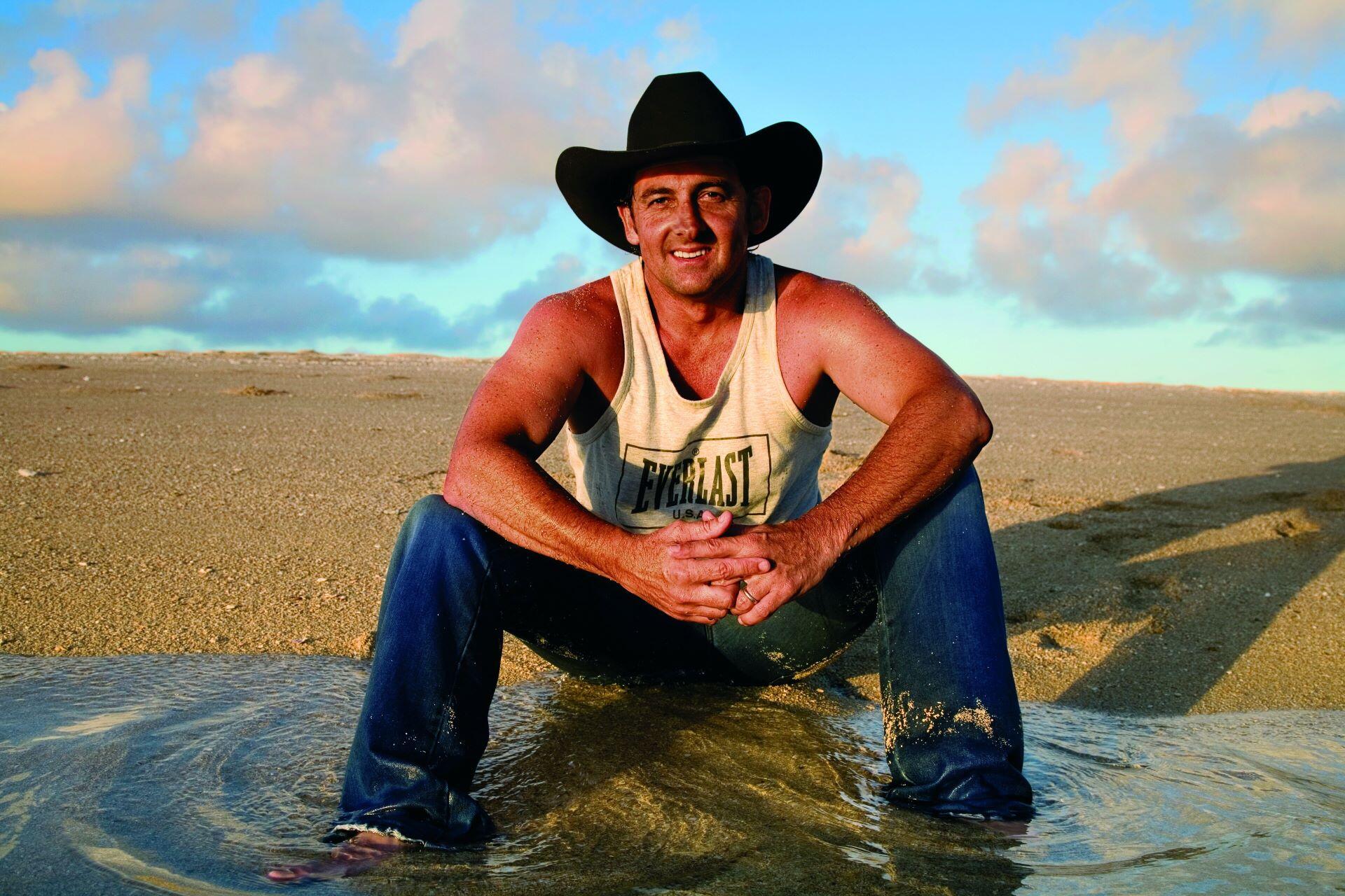 Lee Kernaghan wearing a black cowboy hat while sitting on a beach.