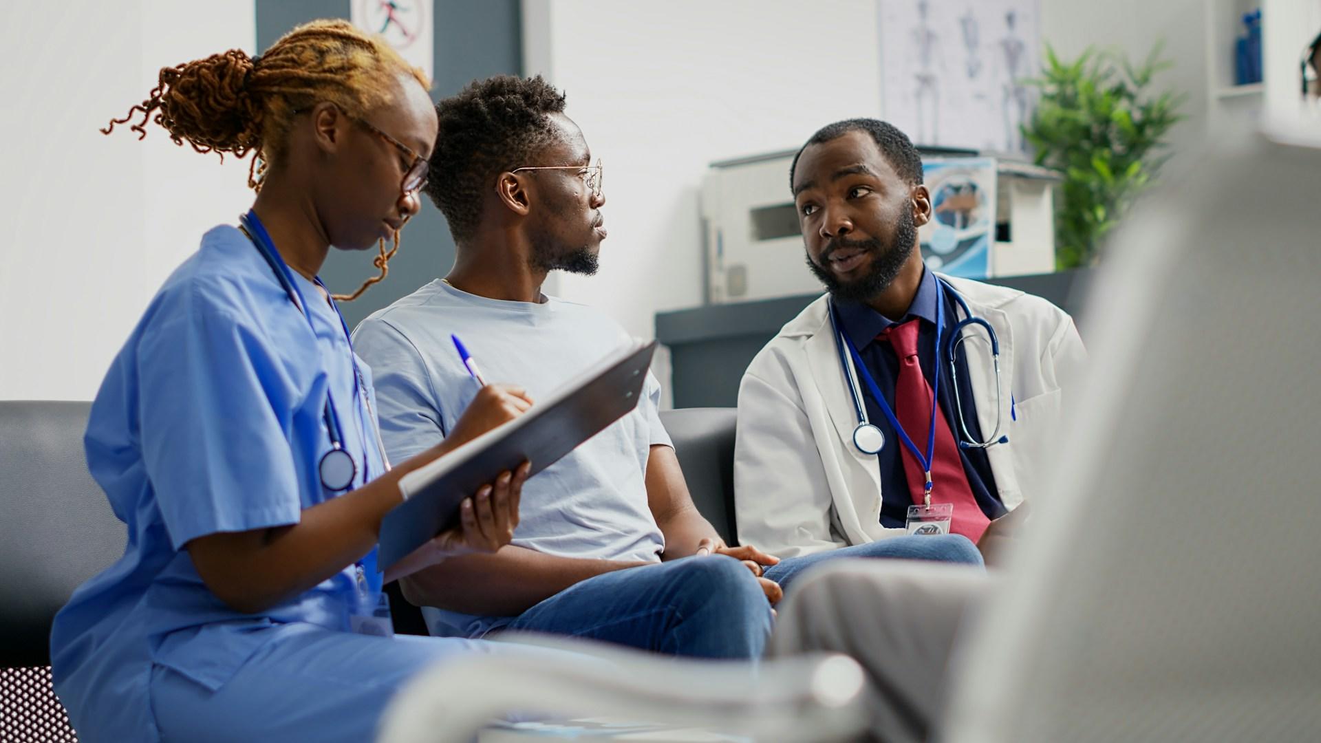 A person in blue scrubs takes notes as a doctor speaks to a patient.