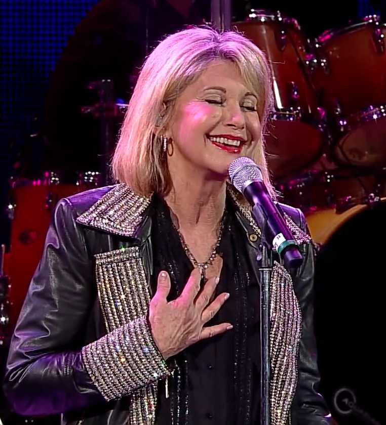 Olivia Newton-John singing on stage wearing a a leather jacket with rhinestones