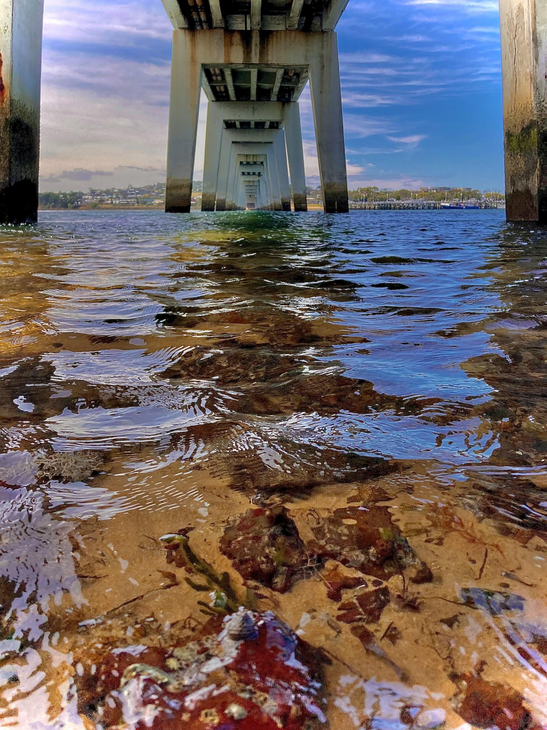Clear water showing red rocks under a bridge. 