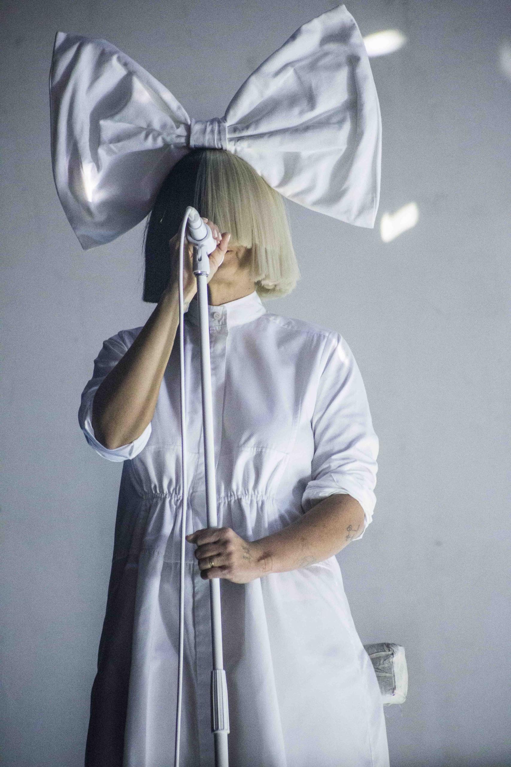 Photo of Sia wearing a white dress and her signature black and blonde wig and large white bow, singing on stage