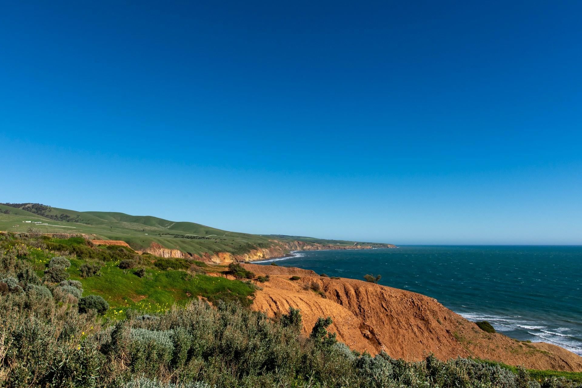 A coastline with green growth and red soil against blue water on a sunny day.