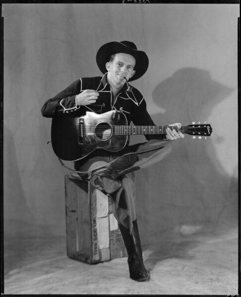 A black and white posed photo of Tex Morton sitting on a box, holding a guitar and a pipe. He is wearing full cowboy garb.