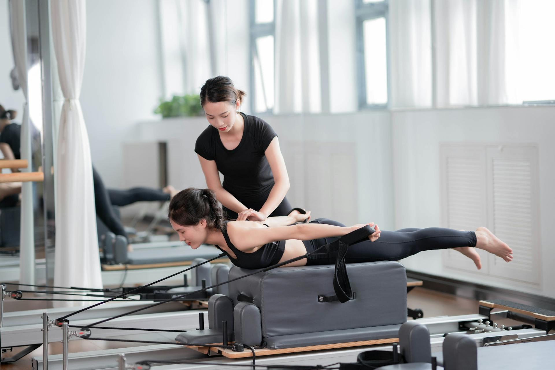 a woman using a Pilates reformer with a female trainer helping her