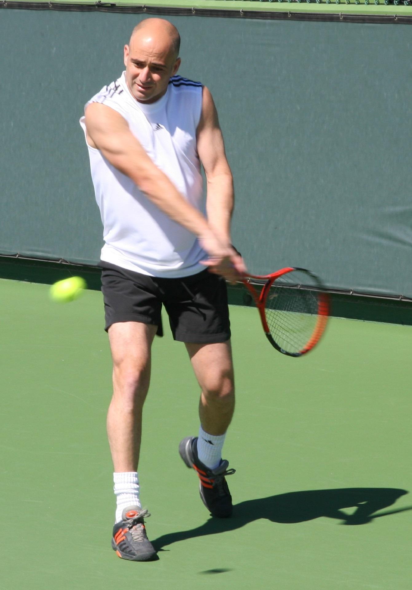 A man in a white shirt and black shorts swings a tennis racket on a grass court on a sunny day.