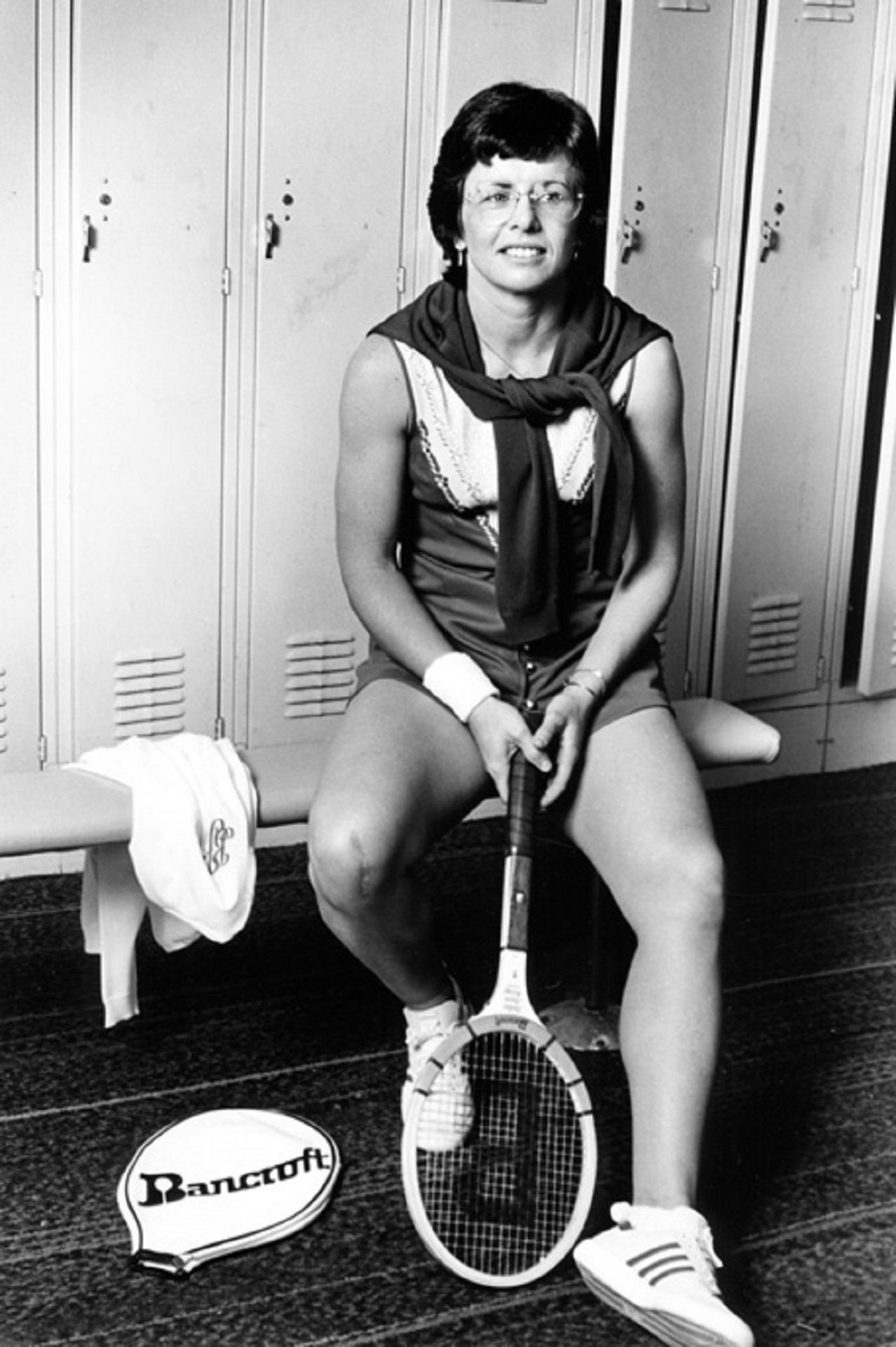 A woman sits on a bench in front of lockers, holding a tennis racket.