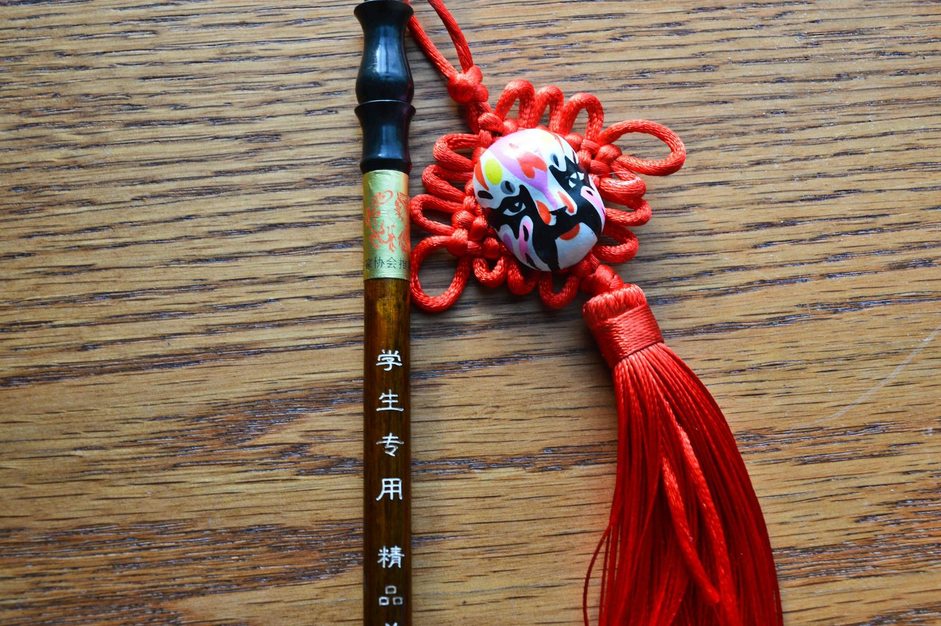Chinese opera mask pendant with tassel alongside a bamboo brush pen on a wooden surface.