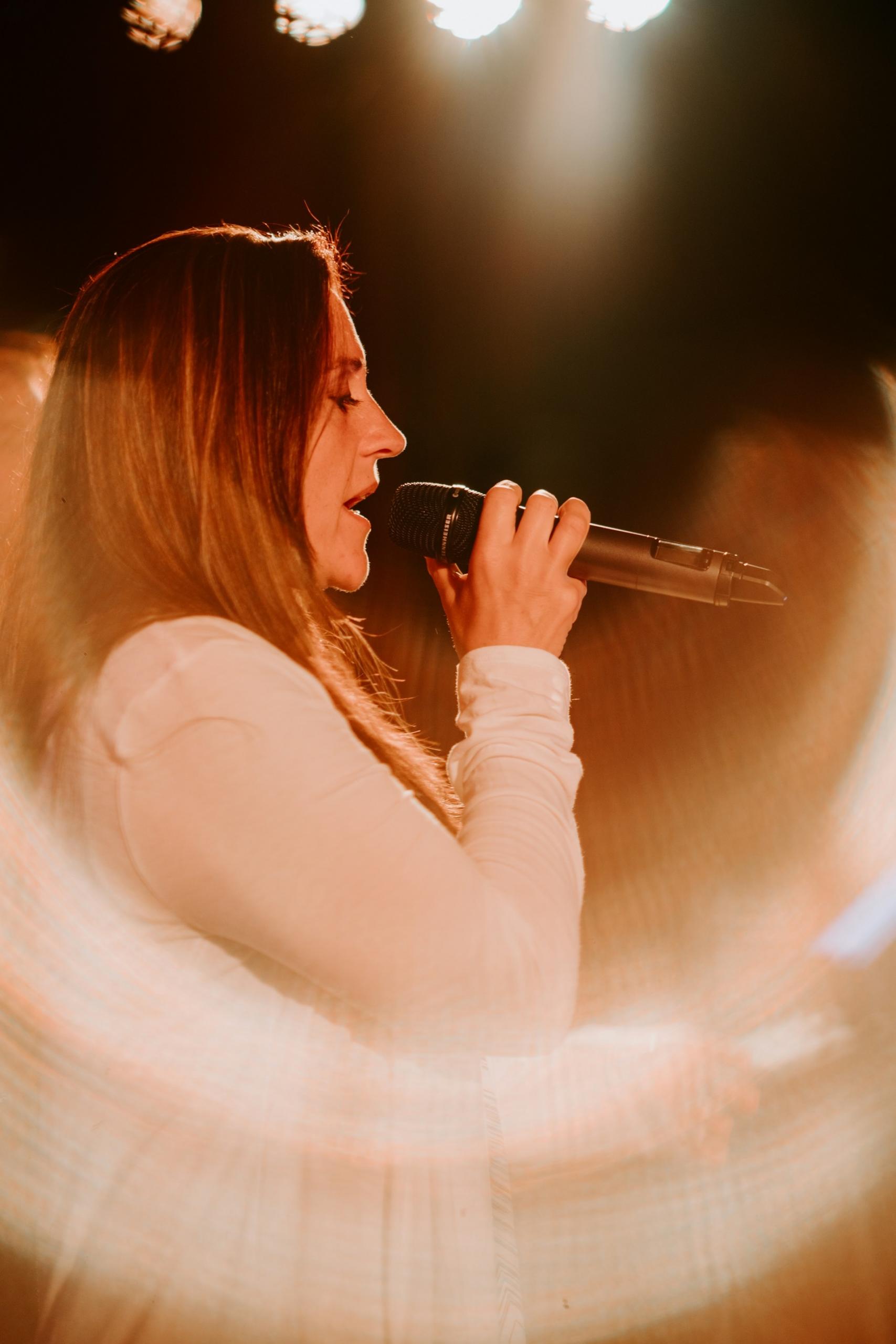 A woman in a white shirt holds a microphone on stage.