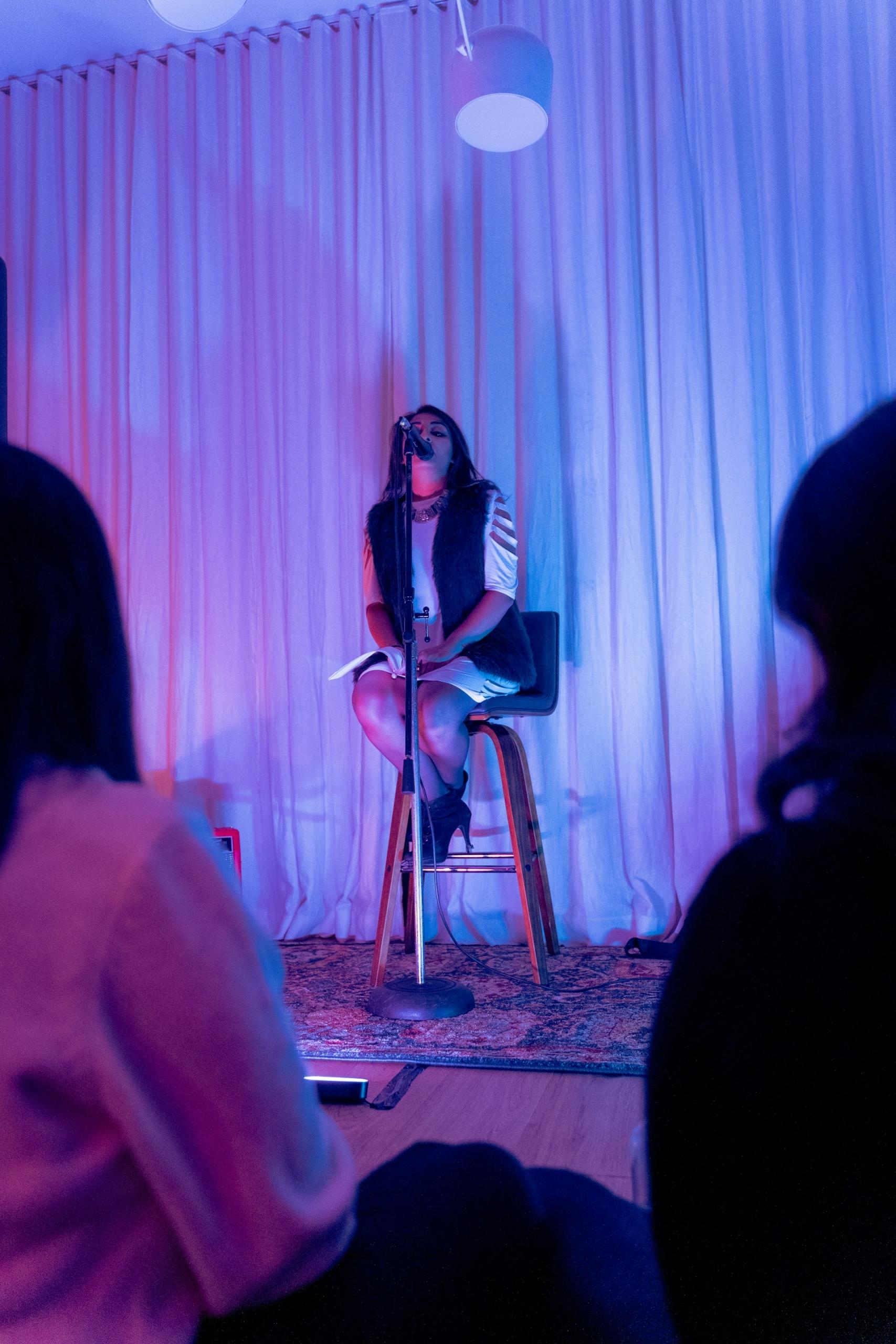 A person sits on a tall stool behind a microphone stand. 