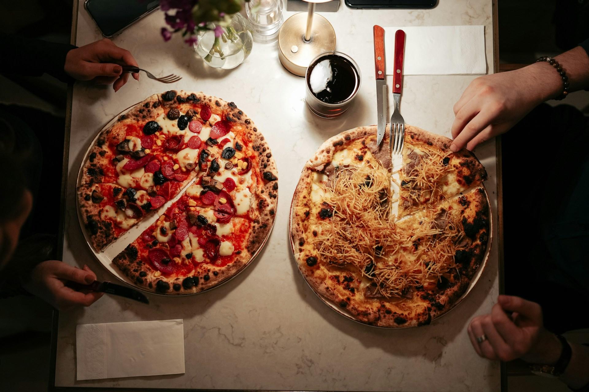 Two people dining with two whole pizzas, one topped with mushrooms and olives, on a table.