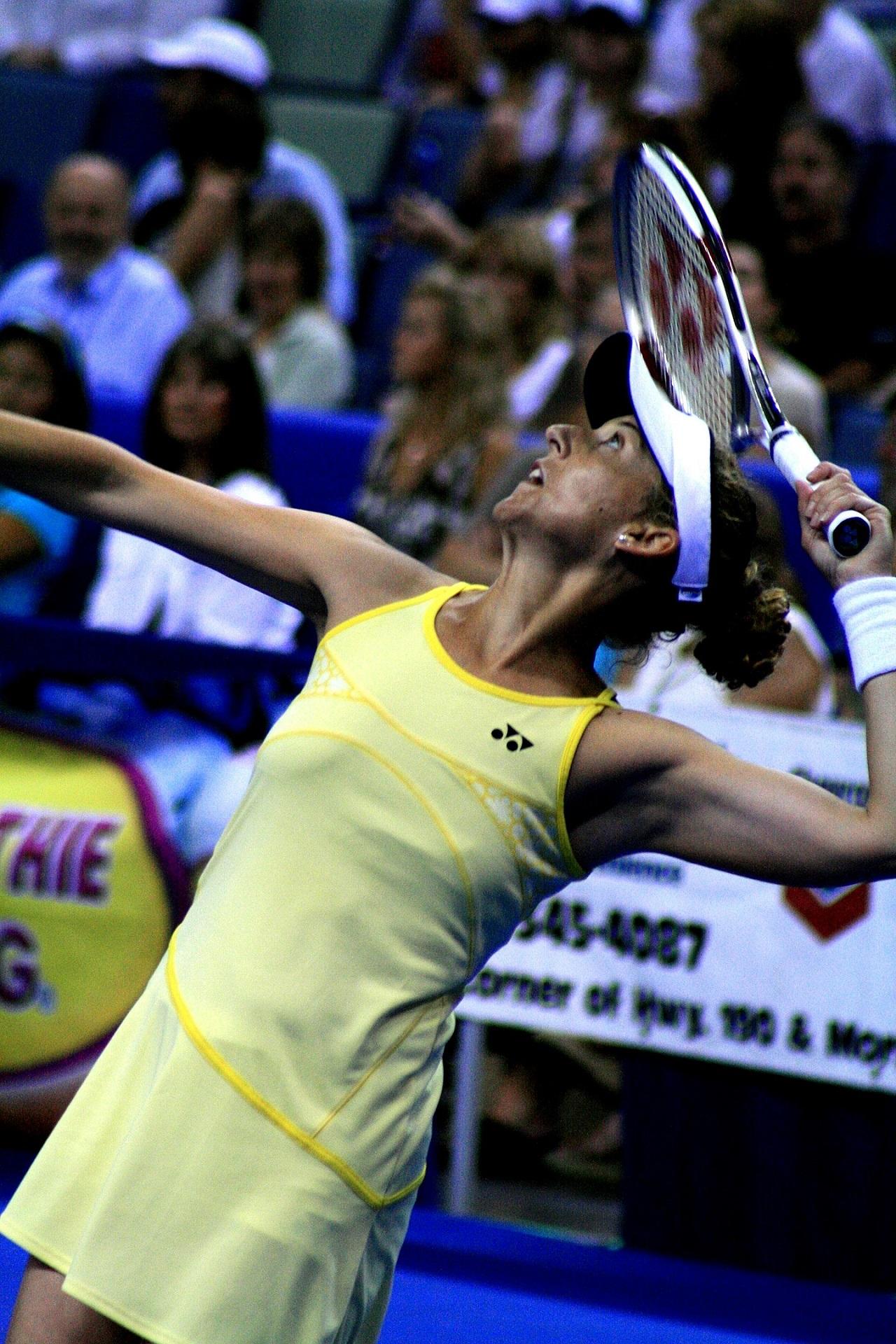 A woman in a yellow dress serves a tennis ball.