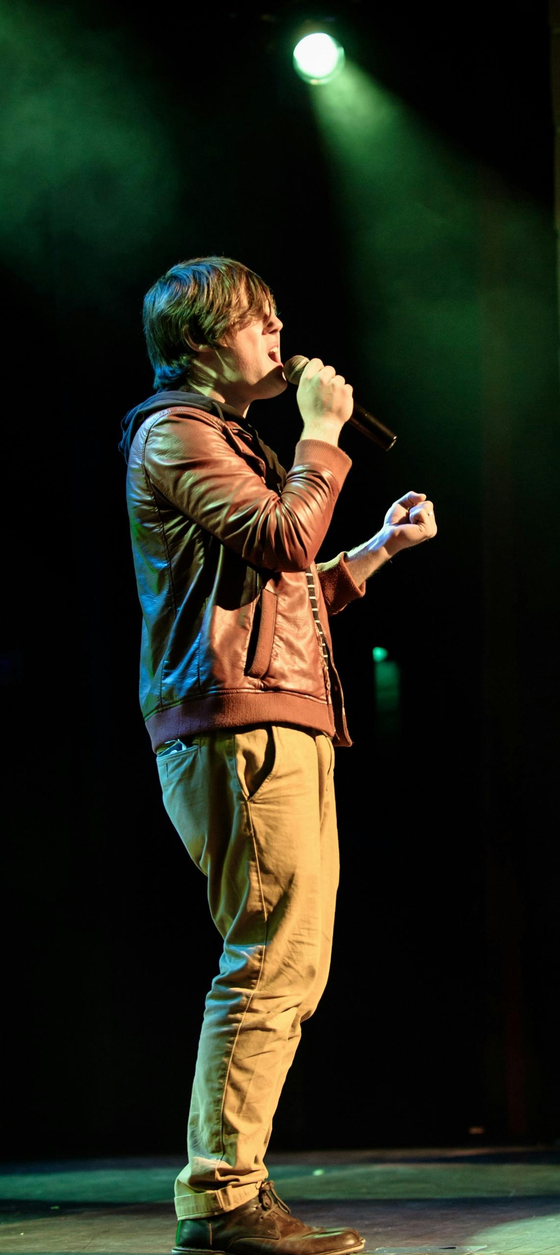 A man in a brown jacket is onstage, holding a microphone.