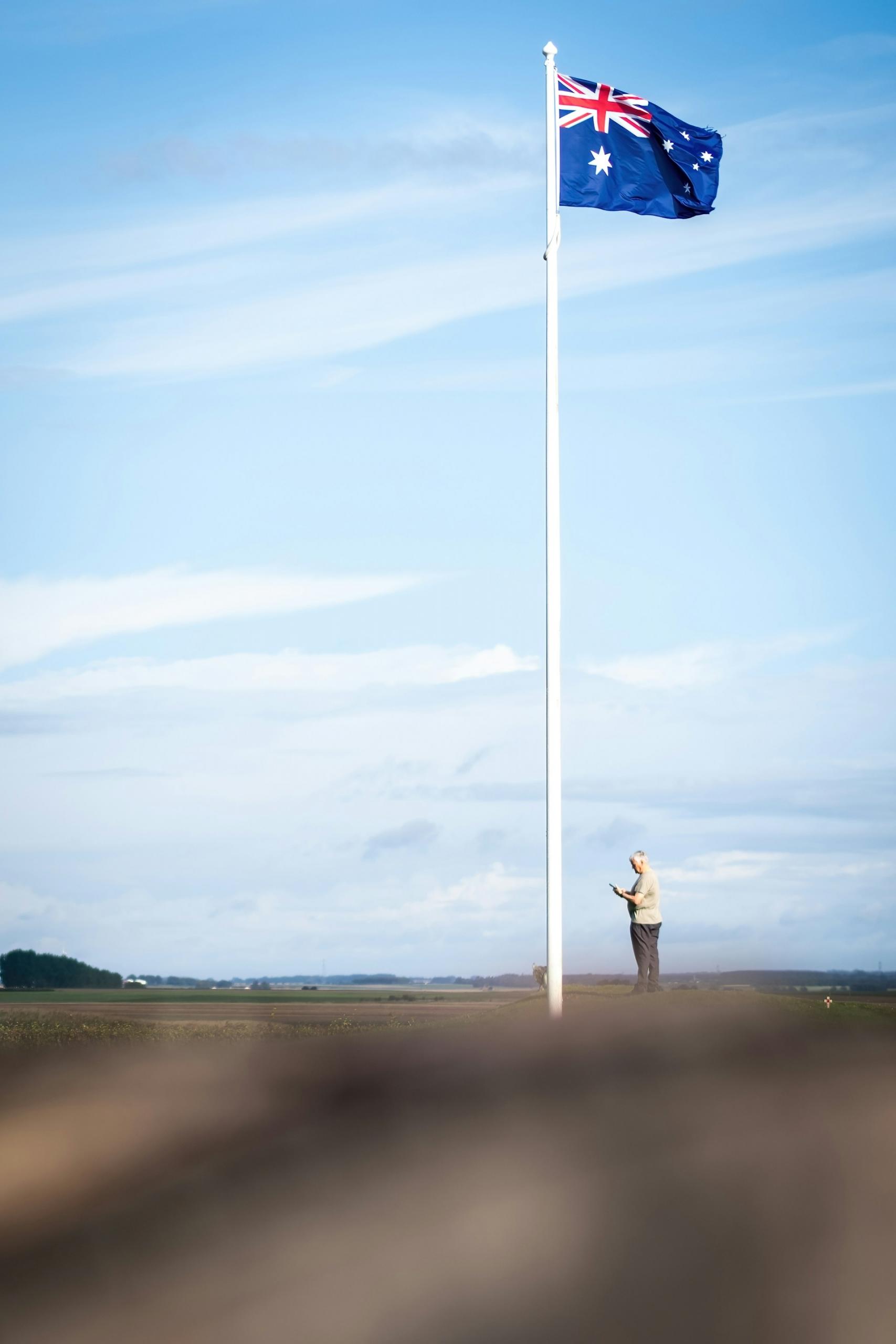 A person stands next to a flag pole flying the Australian flag on a cloudy day. 