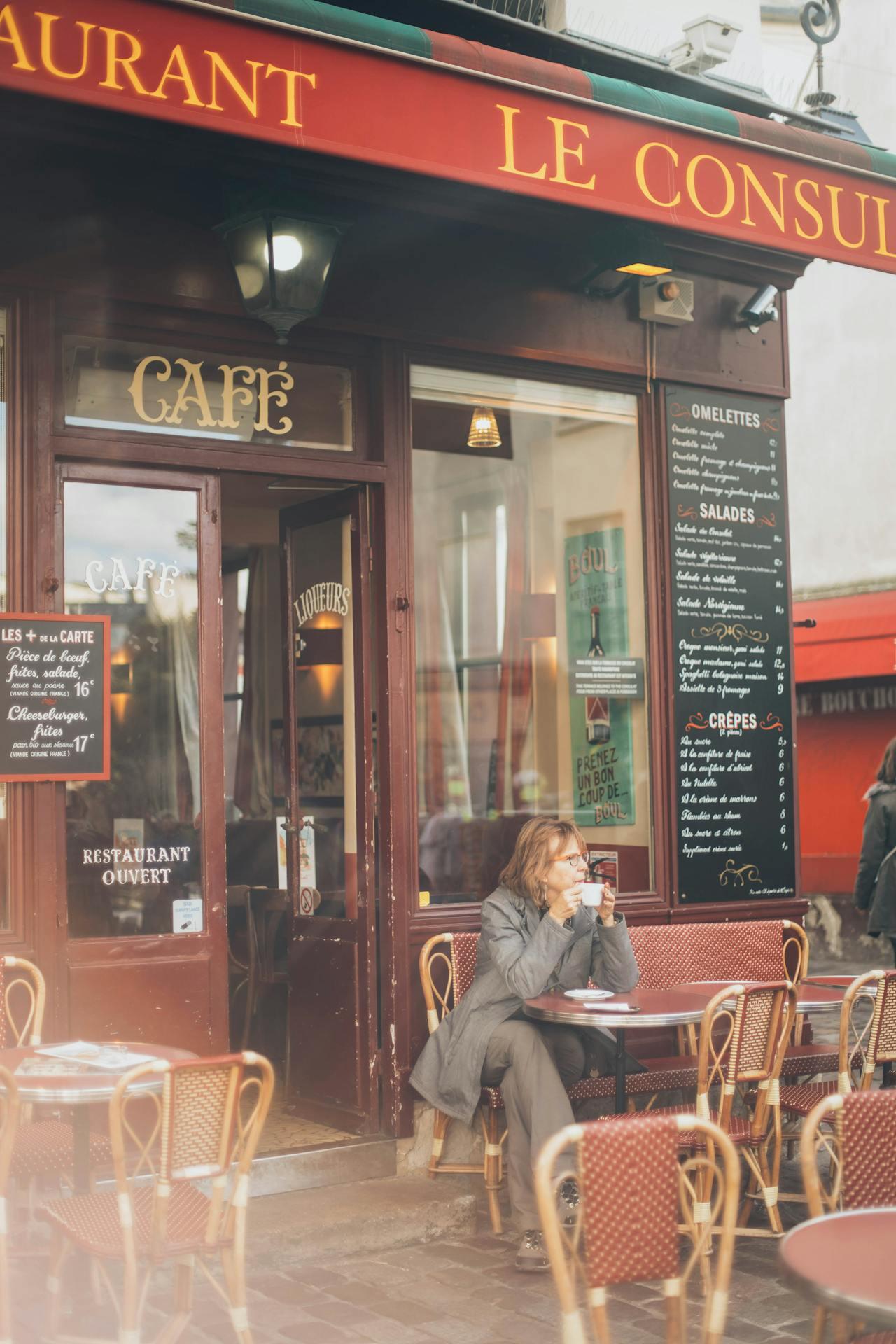 a woman drinking coffee at a table in front of a French cafe