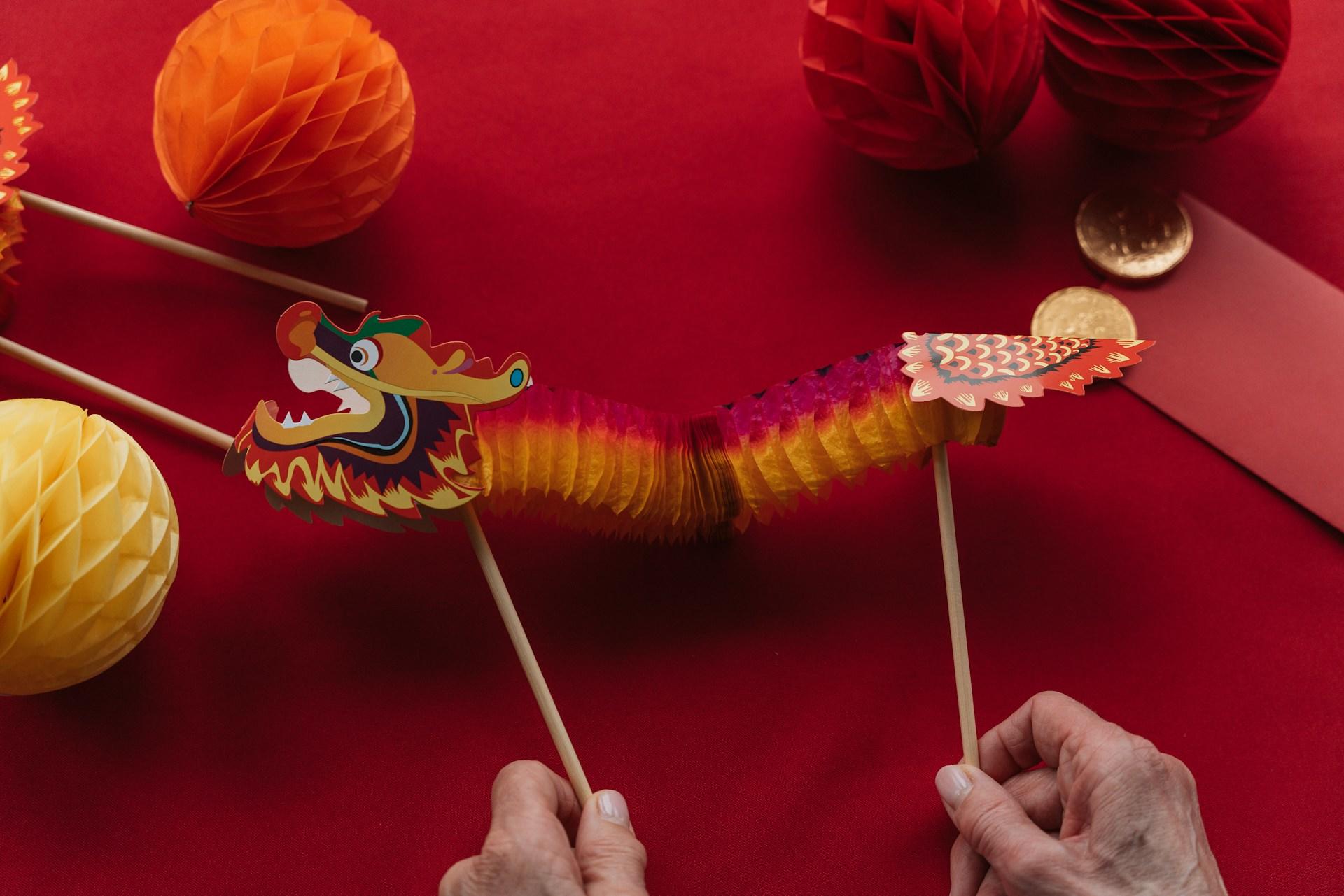 Colorful paper dragon puppet held by two hands against a red background.