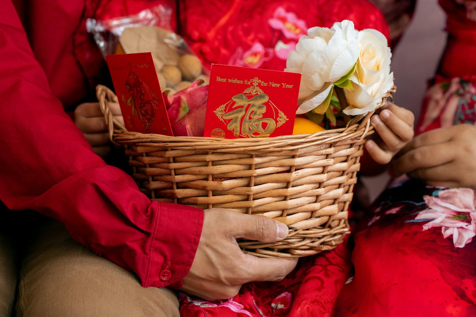 A basket held by people in red outfits, containing fruits, white flowers, and red envelopes.