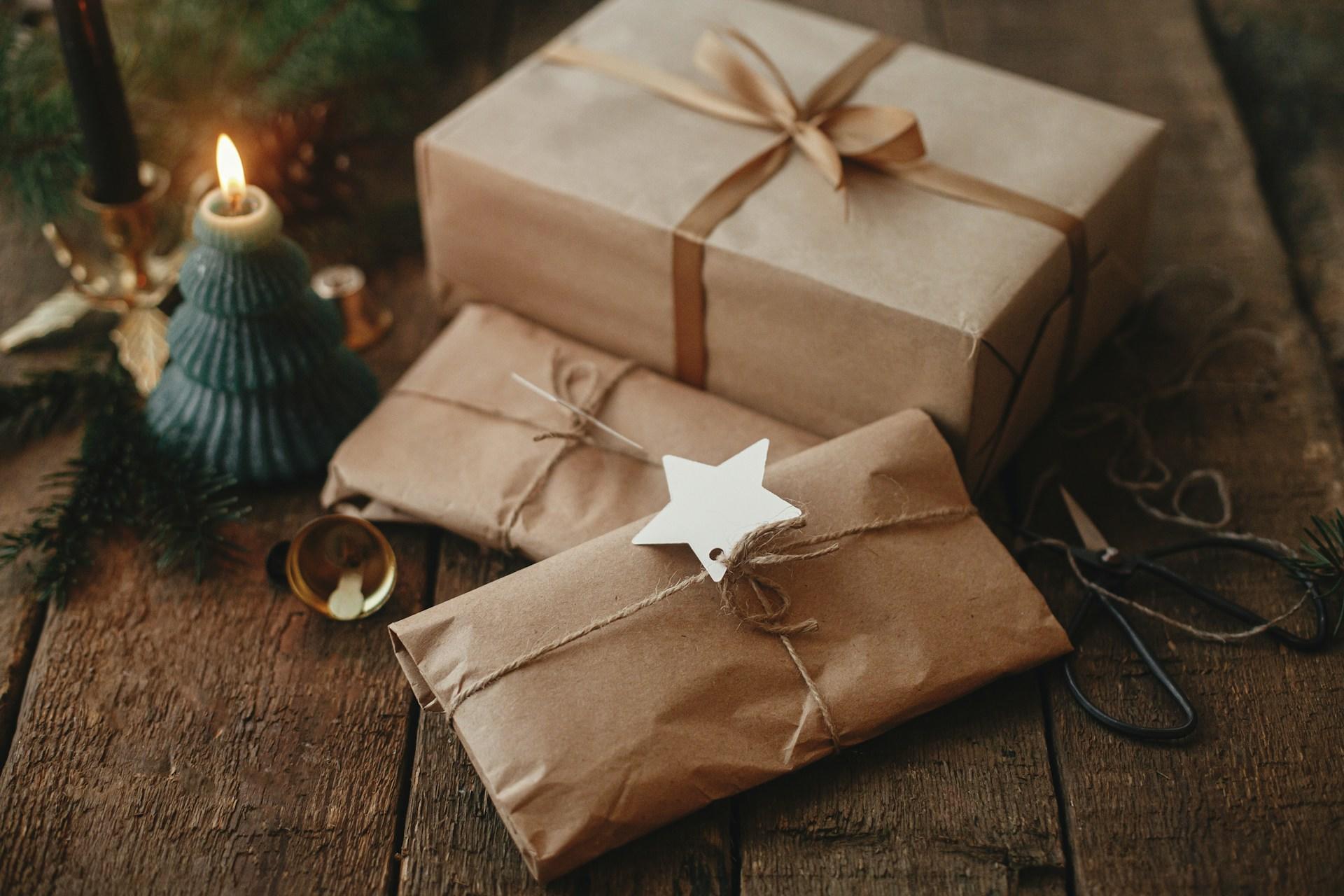 Three wrapped gifts with a lit candle and Christmas decorations on a wooden surface.