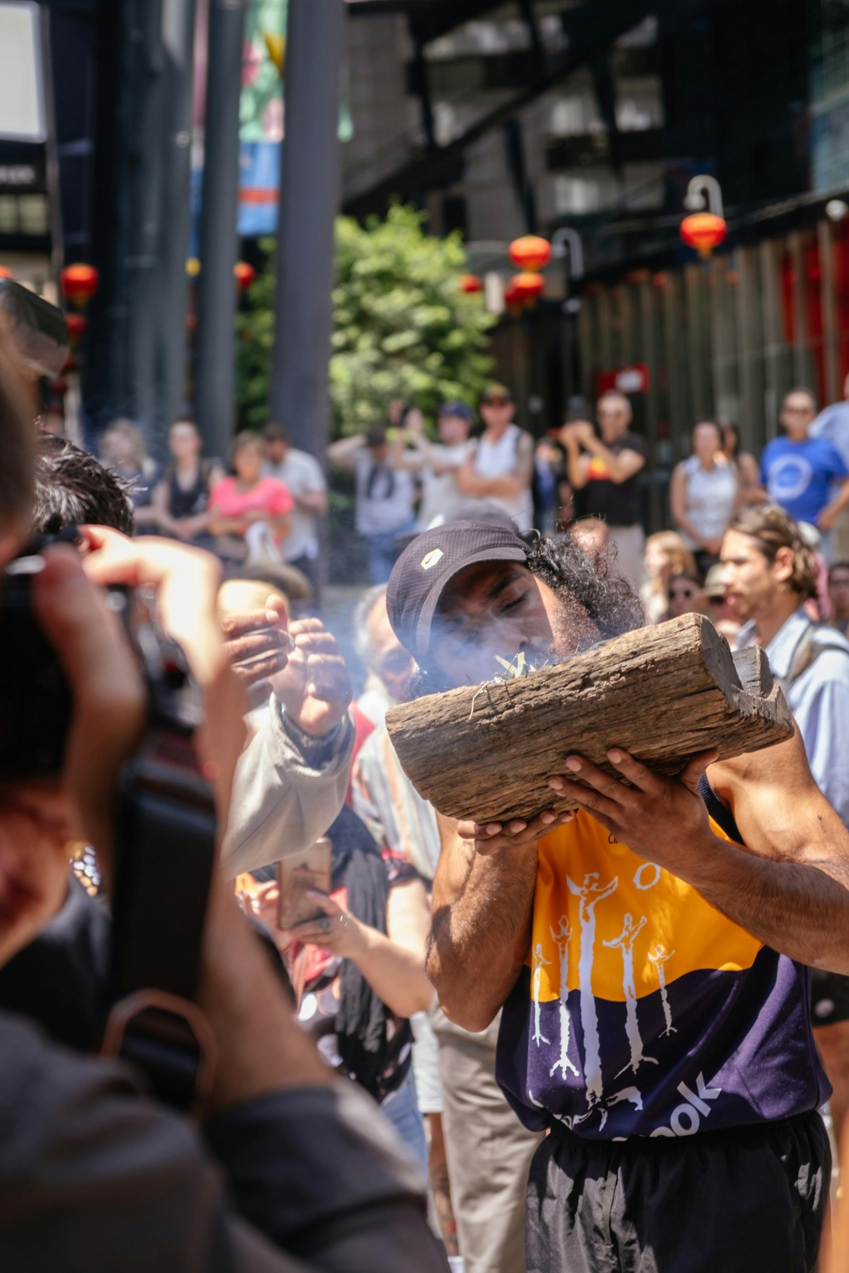 A man holds a burning log in a crowd of people on a sunny day. 