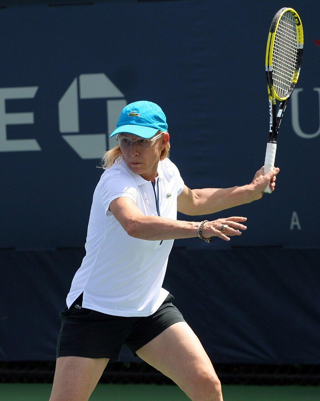A woman in a white shirt and dark shorts plays tennis on a sunny day.