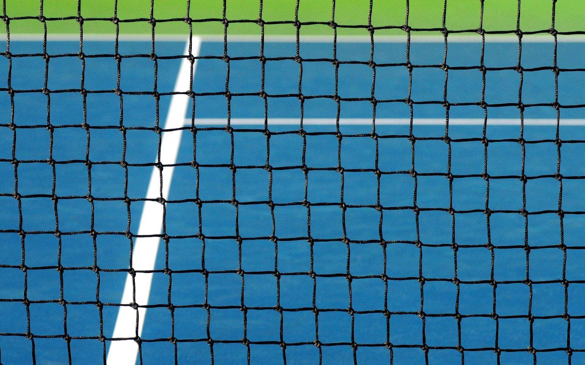 A view of a blue tennis hard court through a net.