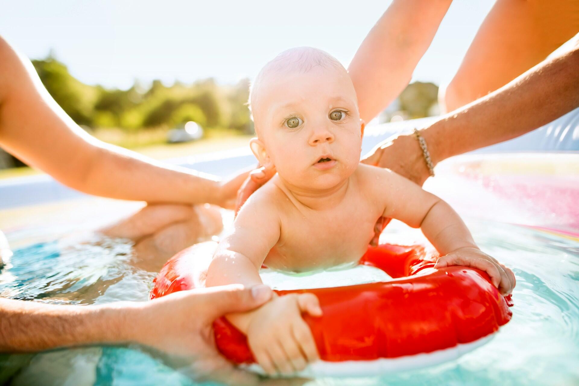 A baby on a red inflatable, with two sets of adult arms supporting it.